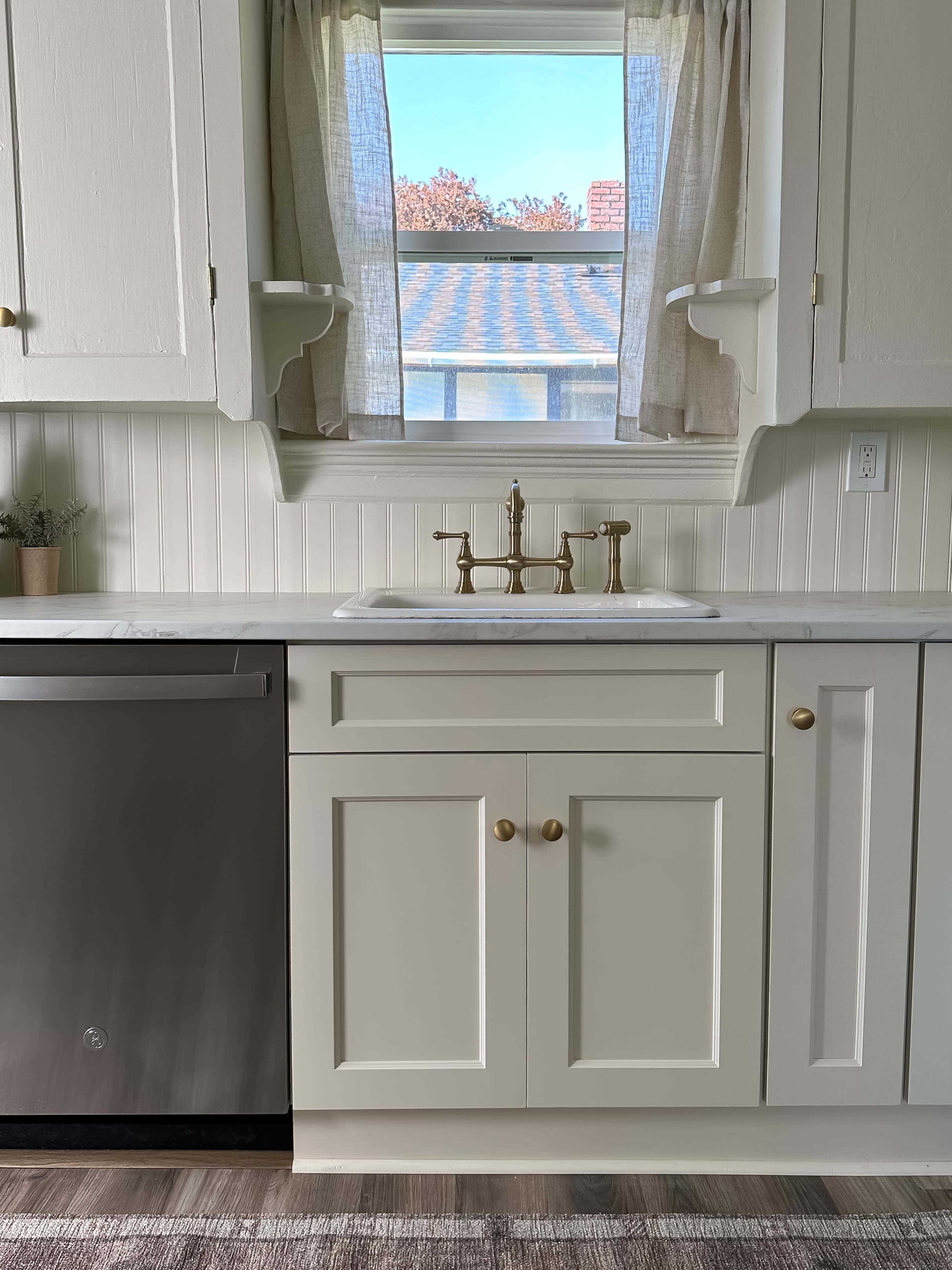 A kitchen area features a sink with a brass faucet, beige cabinets, and a window with curtain overlooking a roof.