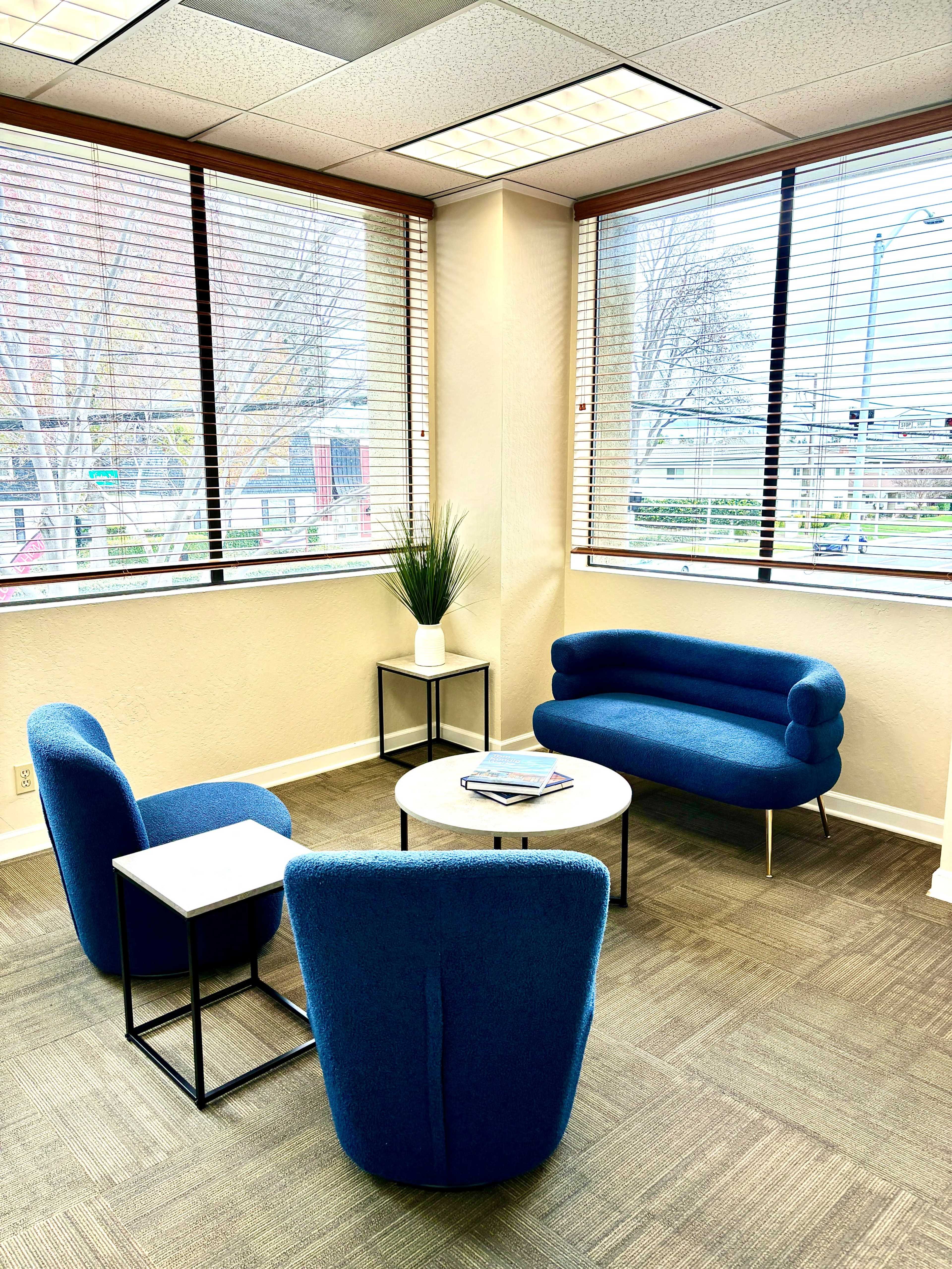 A modern waiting area with blue upholstered chairs and a round coffee table, situated next to large windows with blinds.