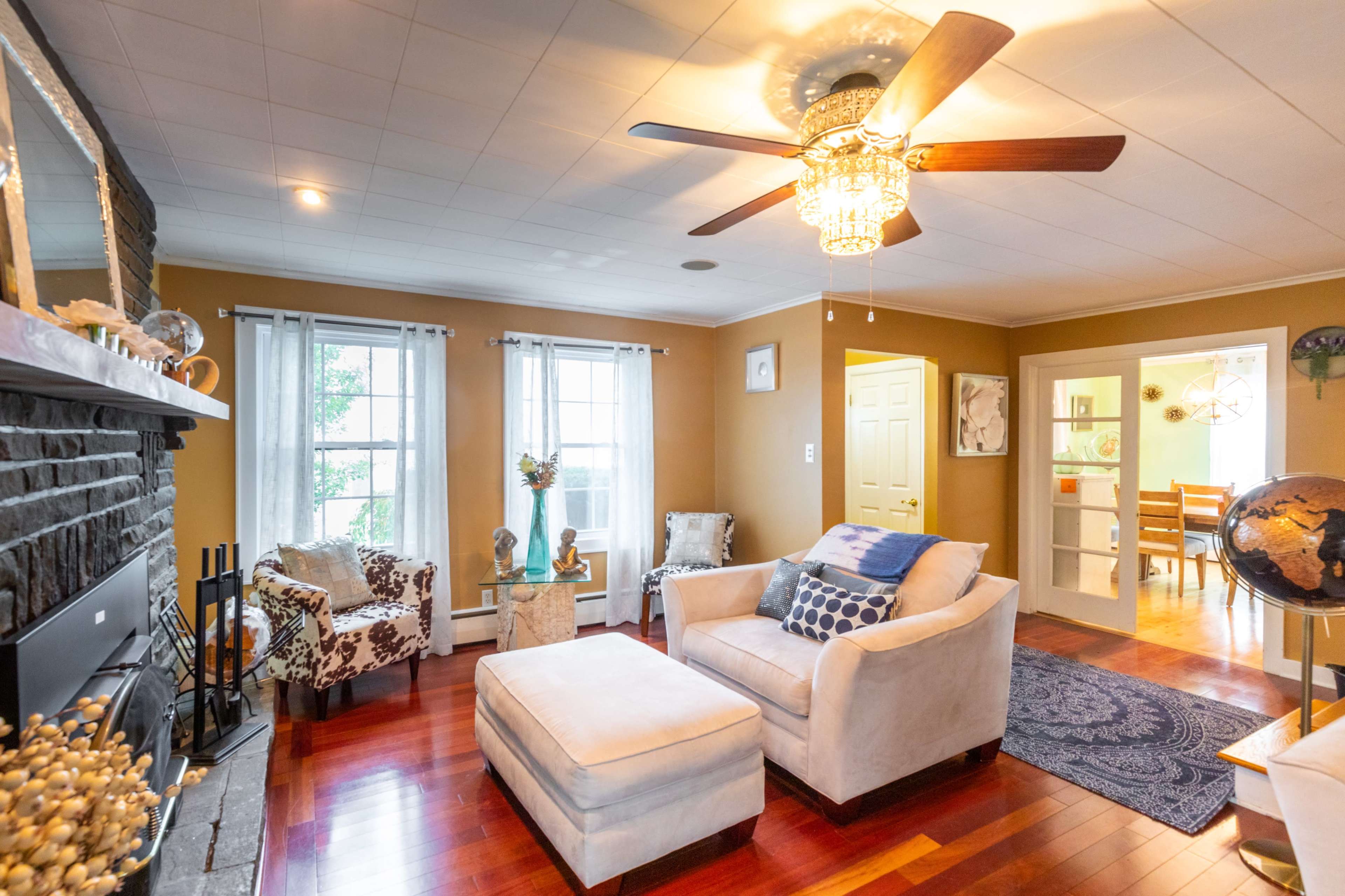 The image shows a cozy living room with a ceiling fan, two chairs, a sofa, and a hardwood floor, complemented by natural light from windows.