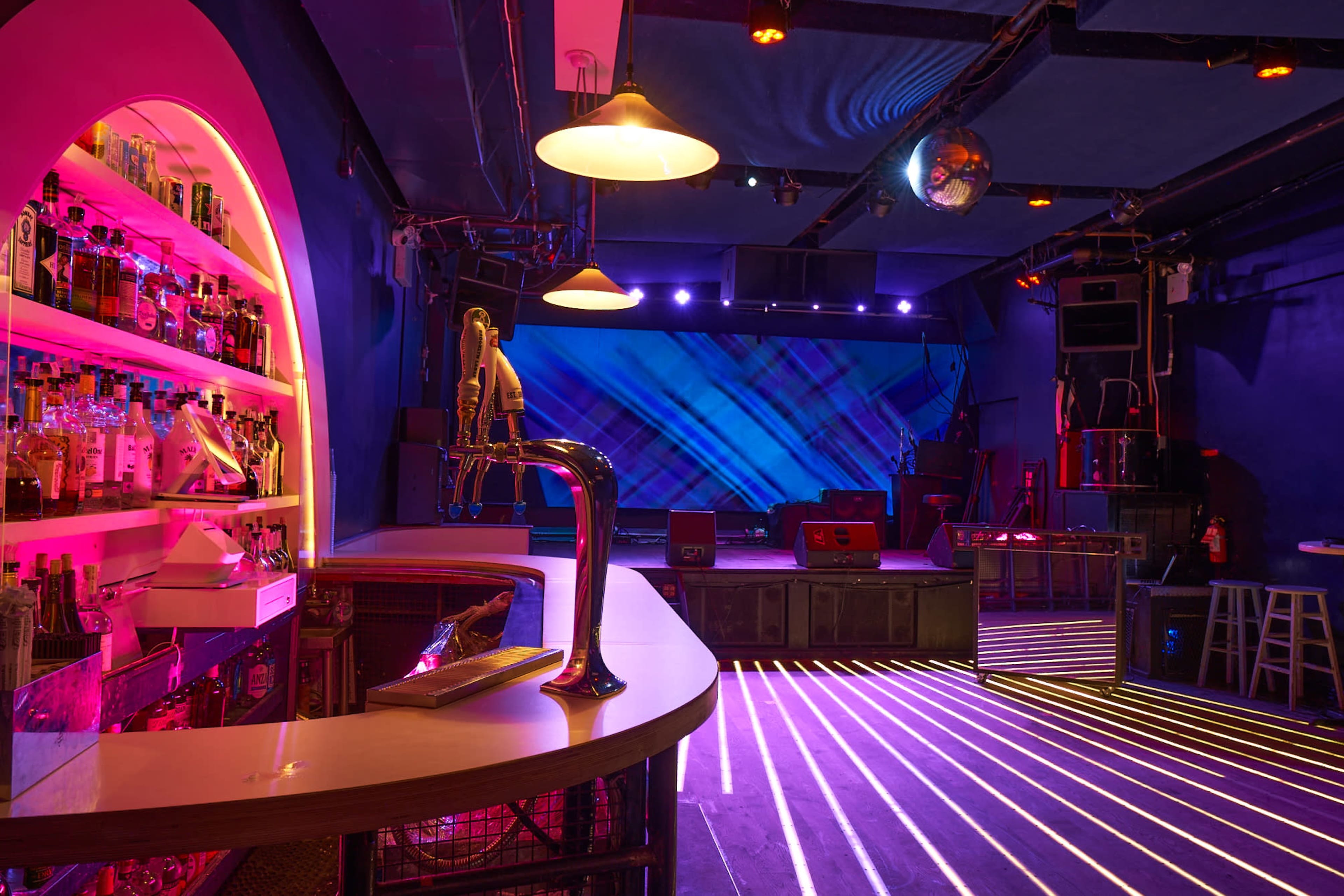 A bar area in a dimly lit venue with a colorful backlit display, shelves of liquor, and stage setup with lighting effects.