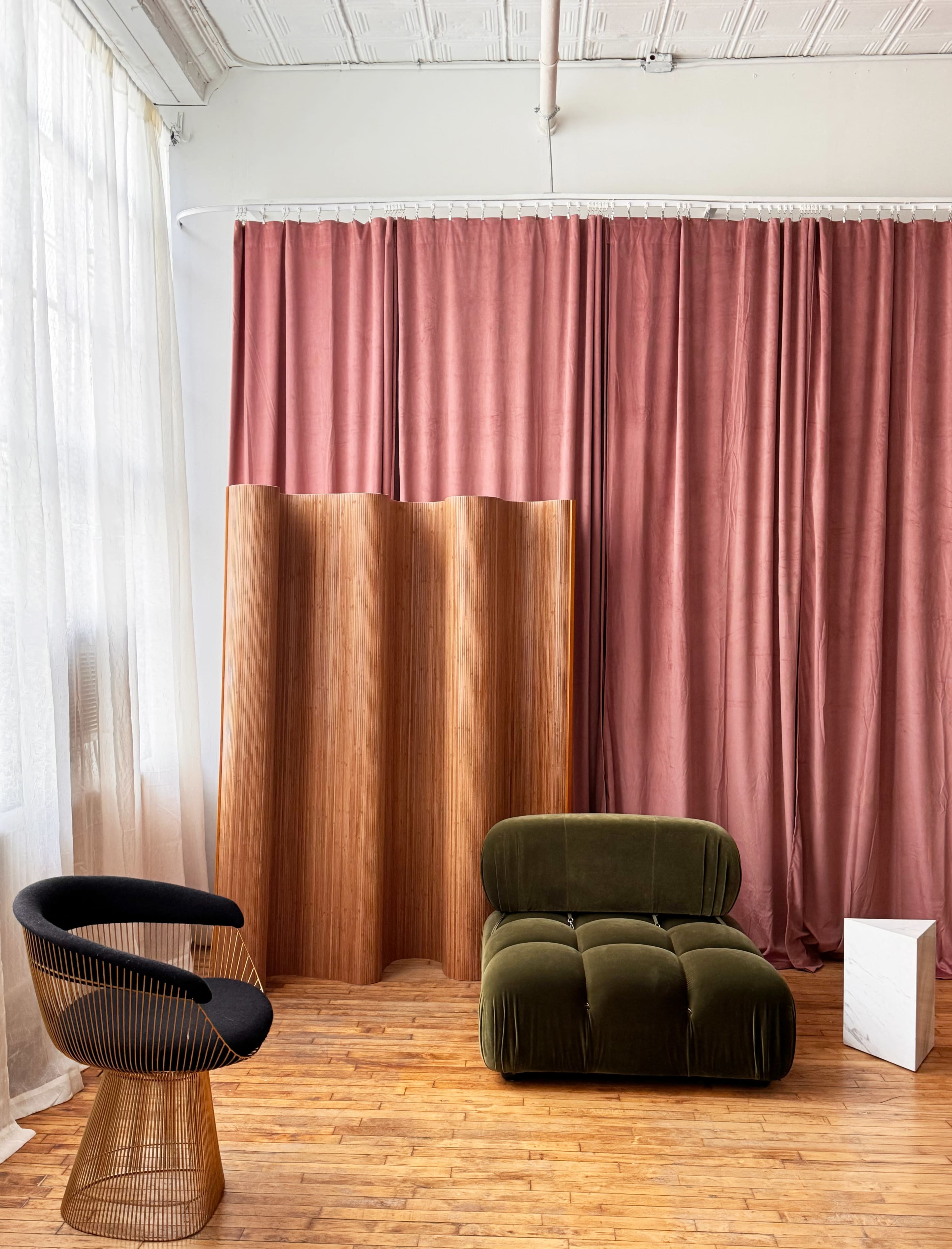 The image shows a cozy interior space with a green velvet chair, a black wire chair, a wooden room divider, and pink curtains, illuminated by natural light from large windows.