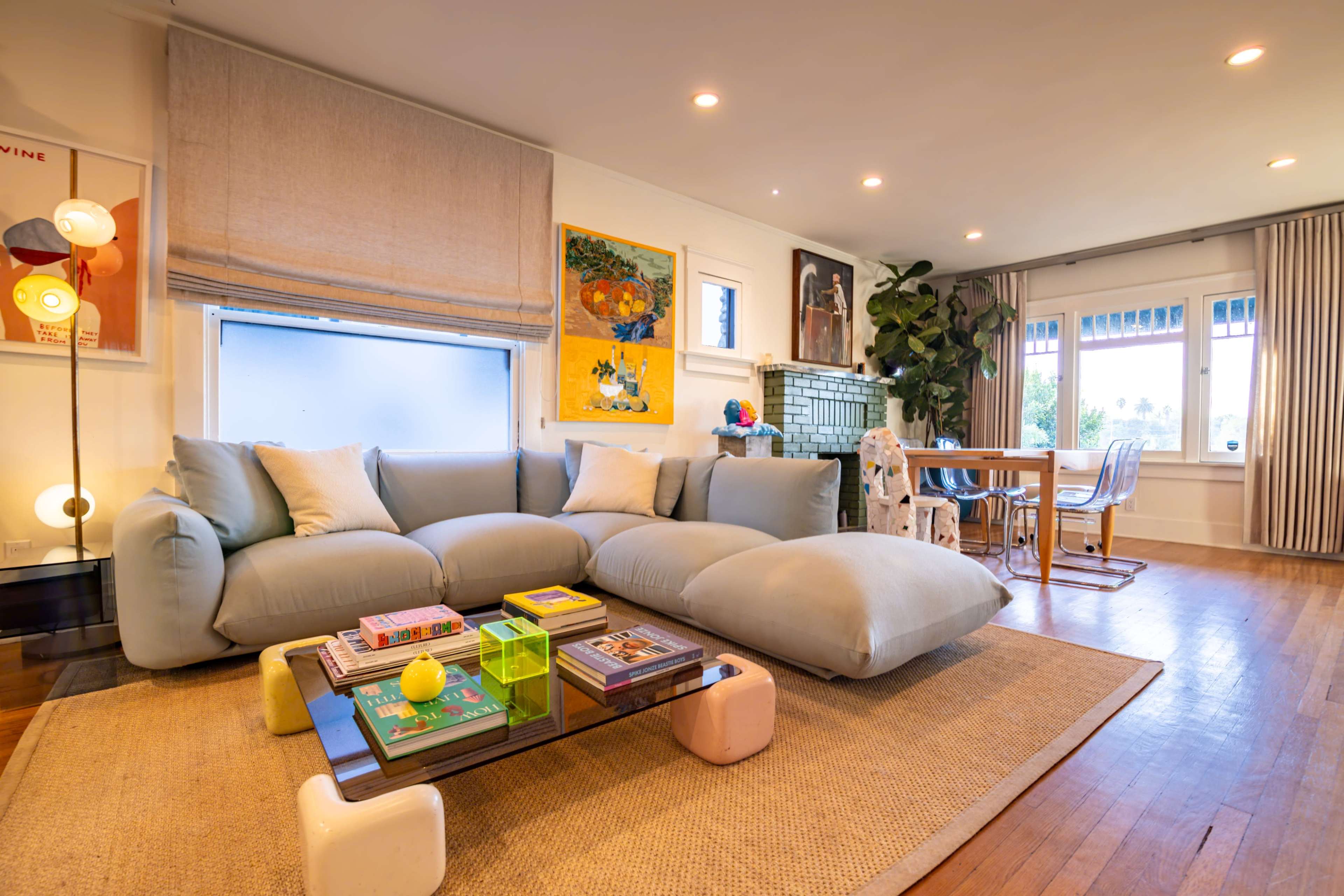 The image depicts a modern living room featuring a light blue sectional sofa, a coffee table with colorful decor, and a dining area with wooden chairs and a table.