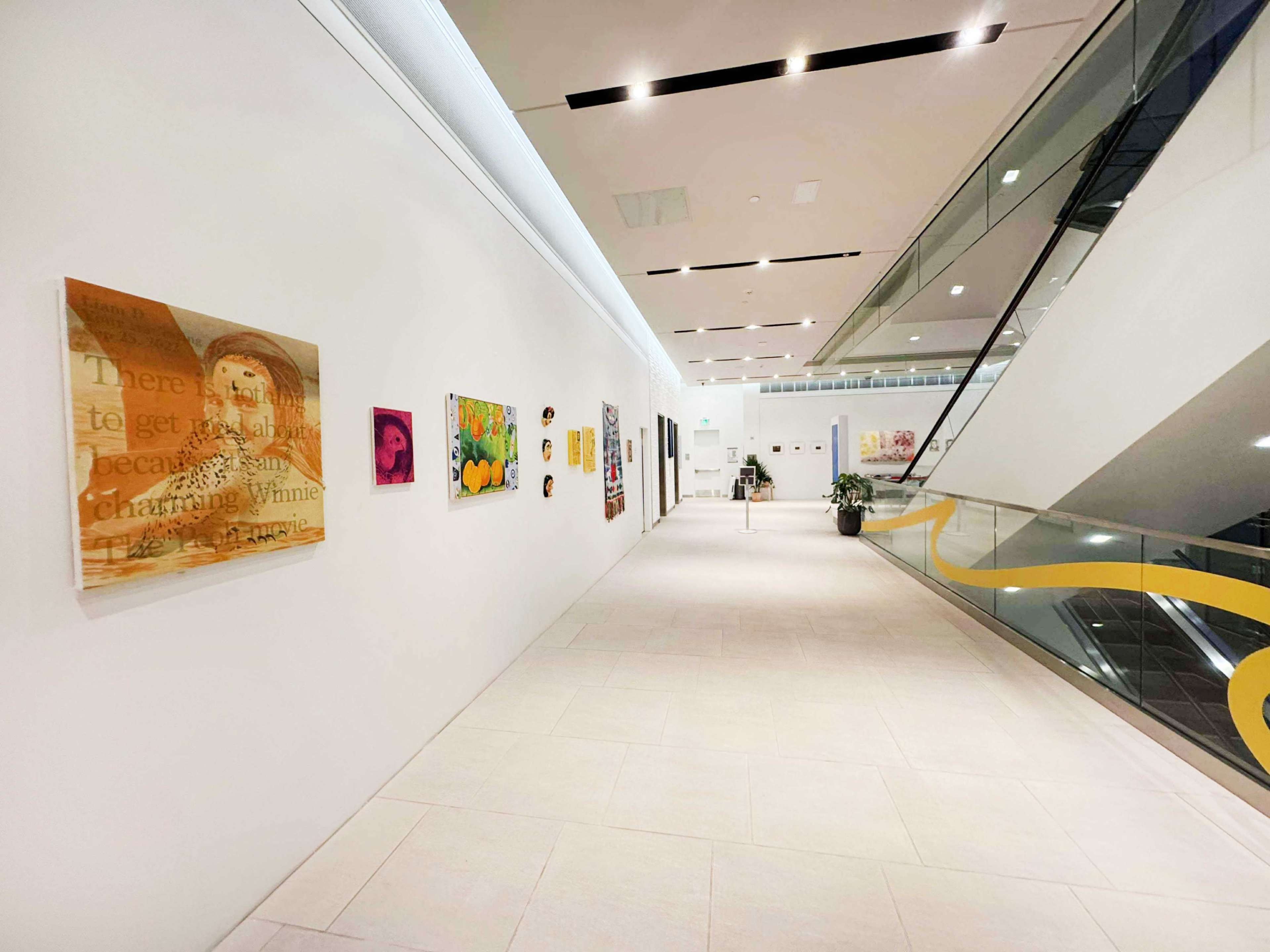 The image shows a modern hallway with white walls displaying various artworks on one side and a glass railing along the staircase on the other.