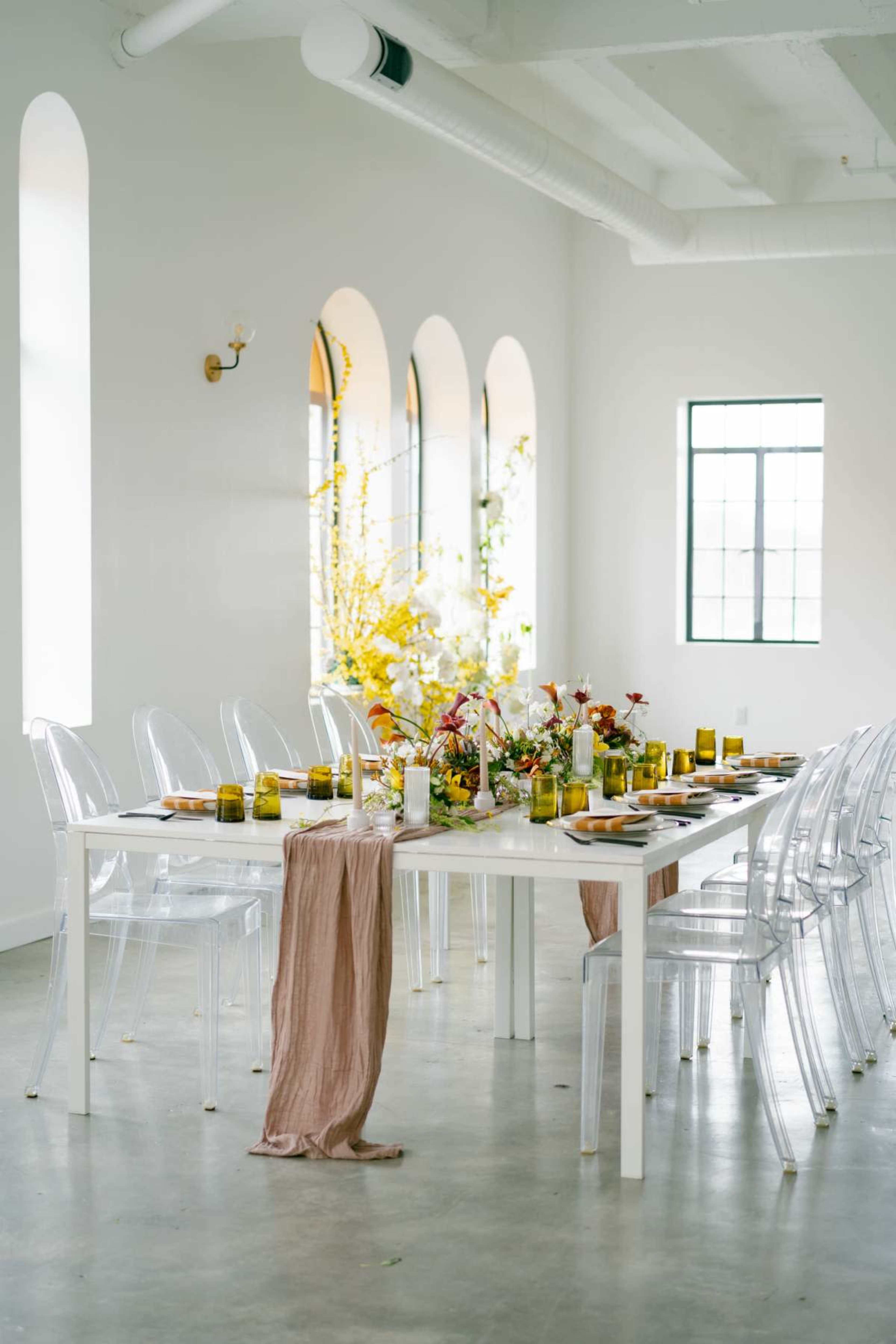 A long white dining table is set with colorful arrangements and glassware in a bright room featuring arched windows.