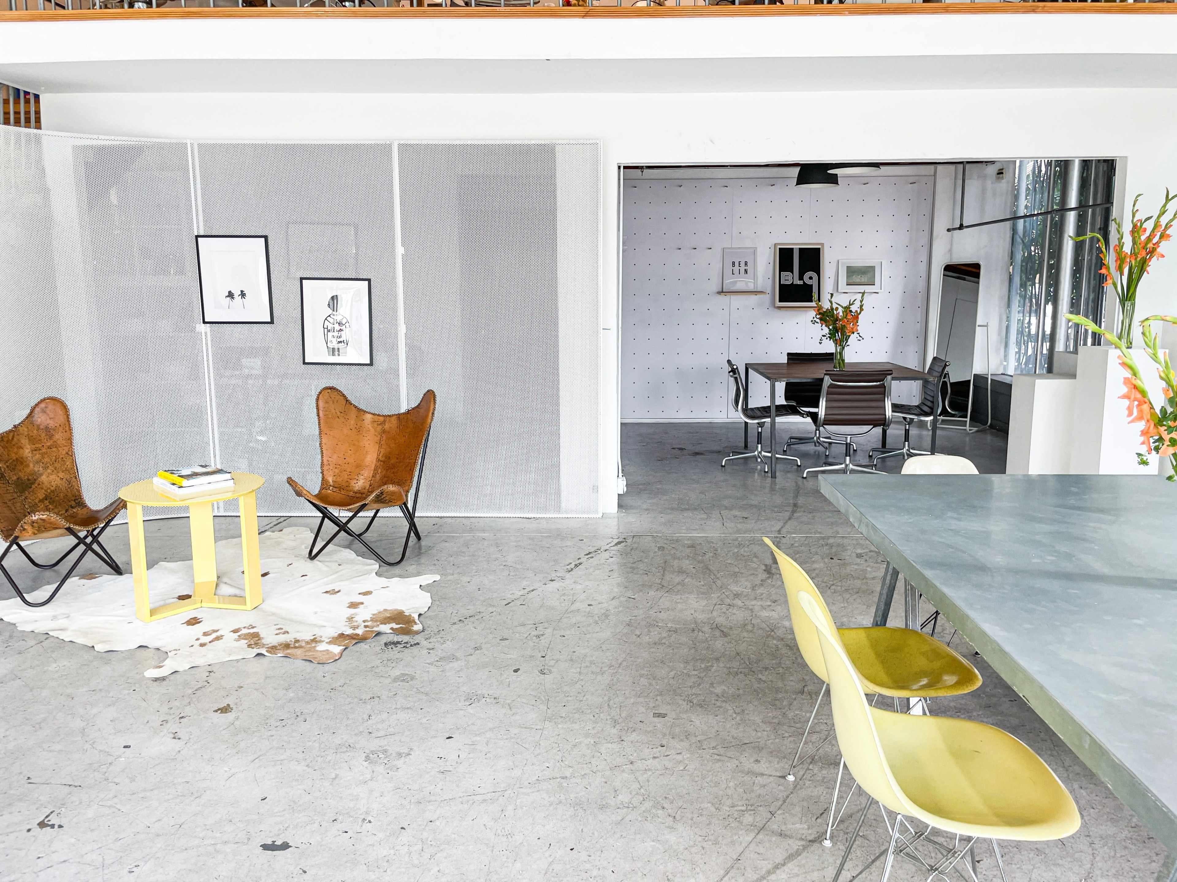 The image shows a spacious interior featuring a seating area with butterfly chairs and a small table, leading to a dining space with a metal table and chairs, separated by a mesh partition.