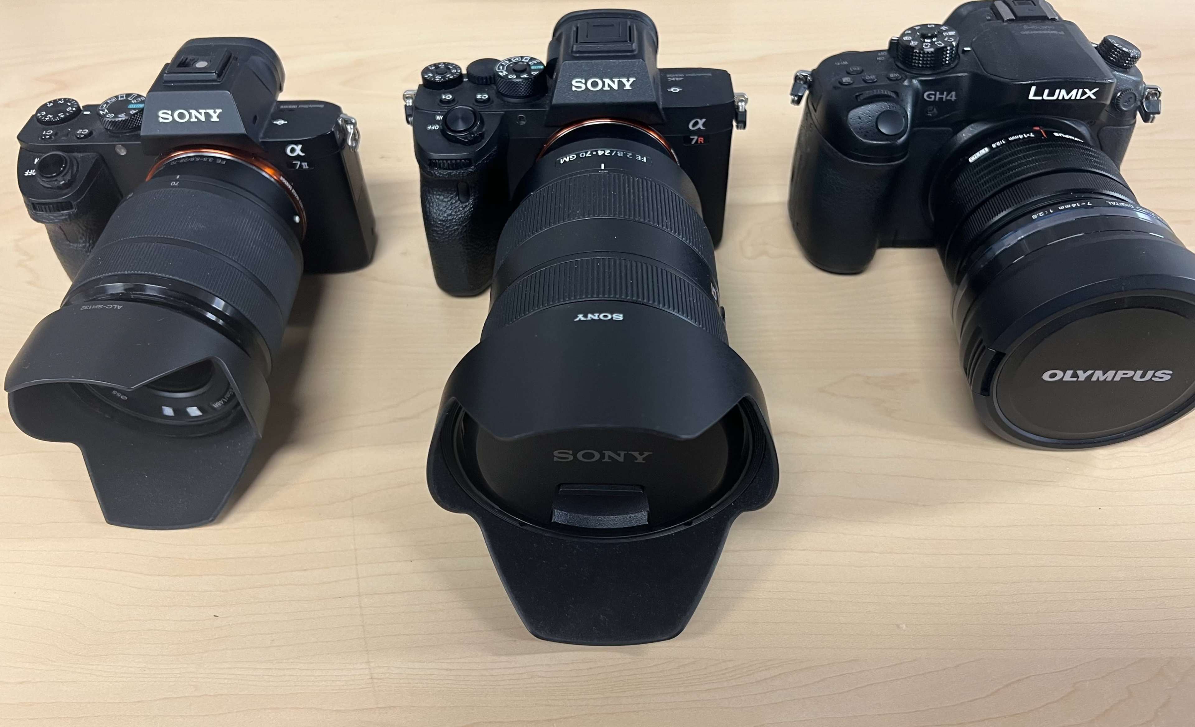 The image shows three cameras lined up on a wooden surface, including two Sony cameras on the left and center, and a Panasonic Lumix camera on the right.