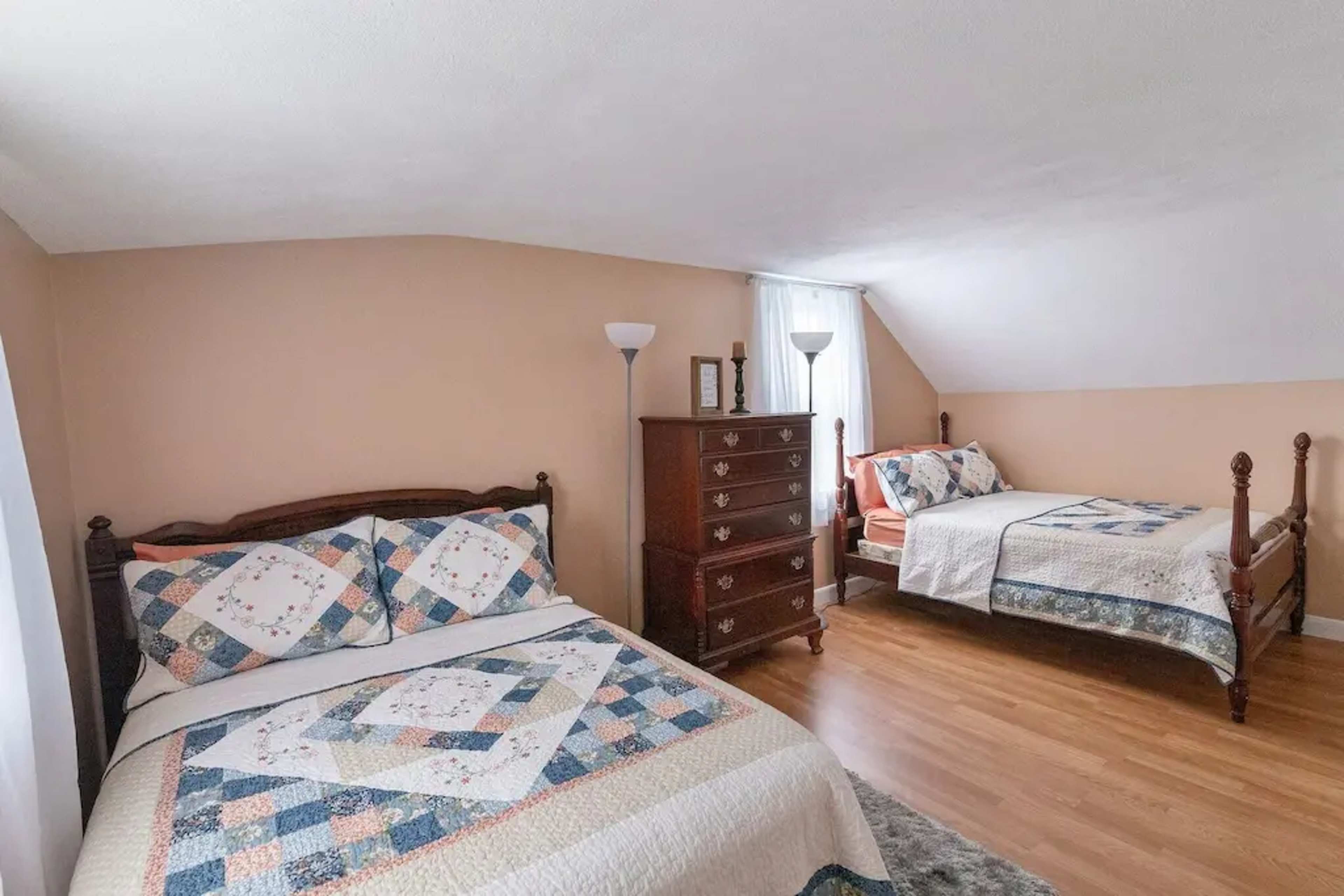A bedroom with two beds, one larger and one smaller, accompanied by a brown dresser and natural light coming through a window.
