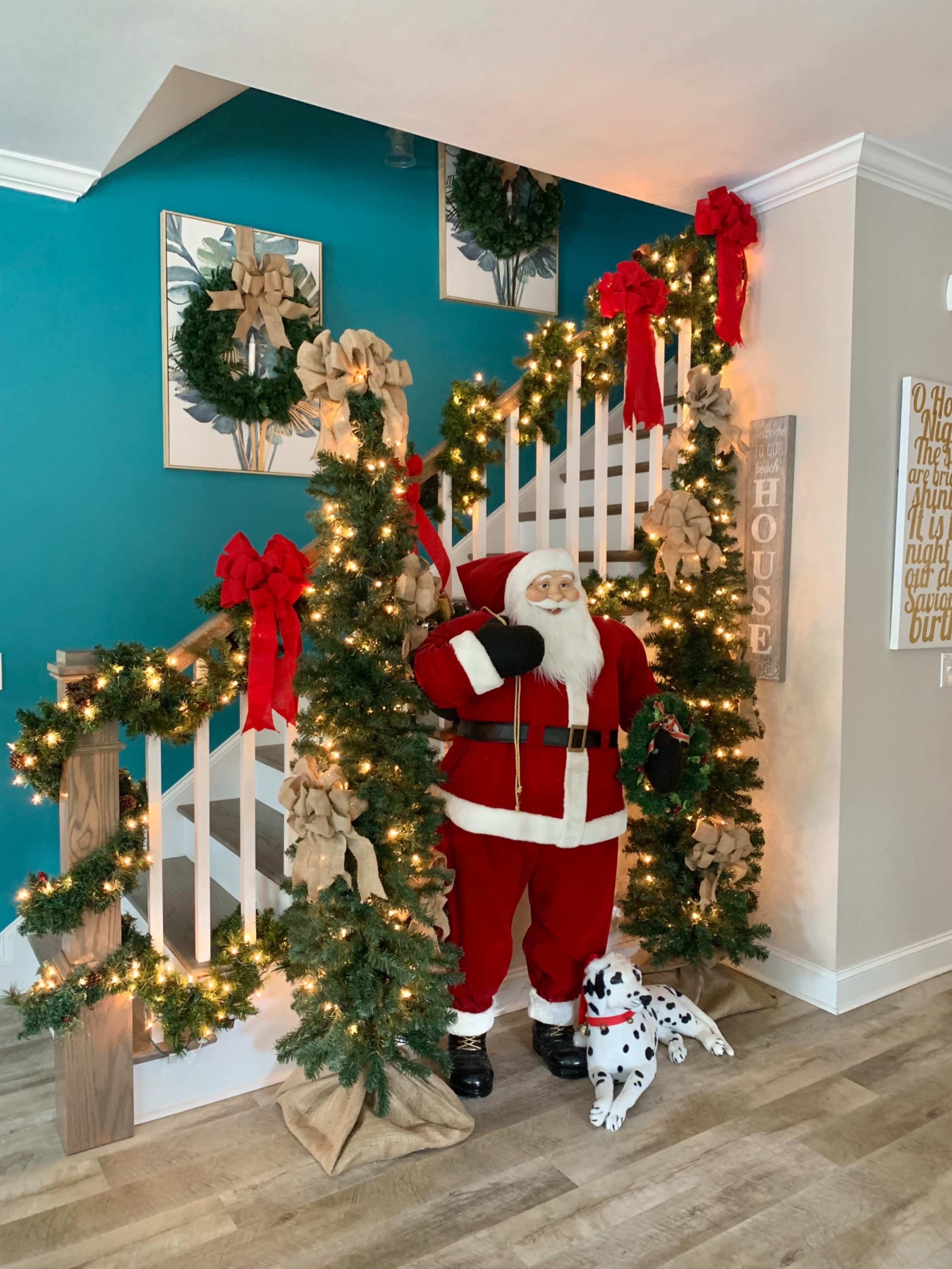 A staircase decorated with garlands, bows, and lights, featuring a life-sized Santa Claus figure alongside a Dalmatian statue.