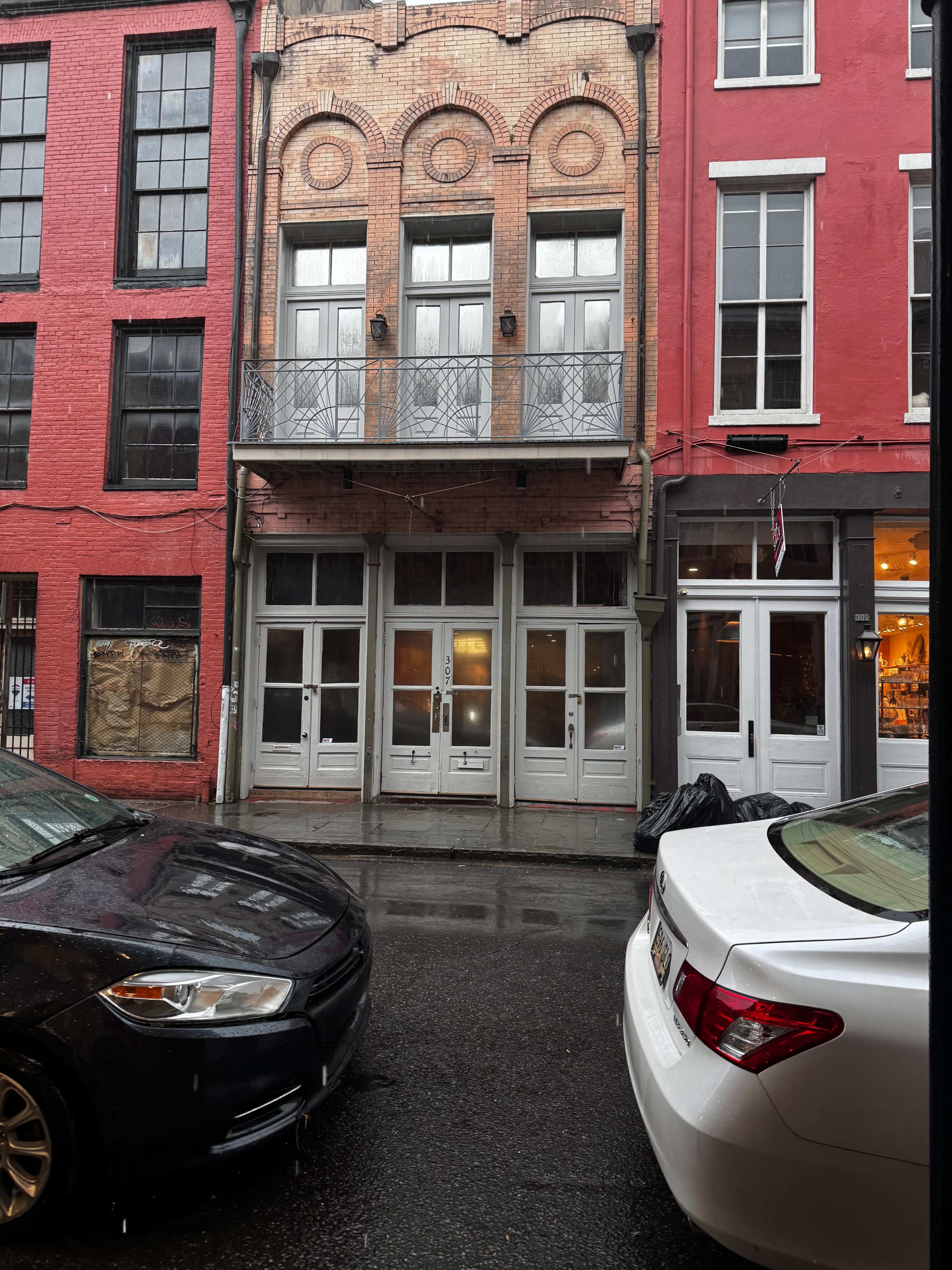 The image shows a street scene featuring a three-story building with red and brick facades, flanked by parked vehicles on a rainy day.
