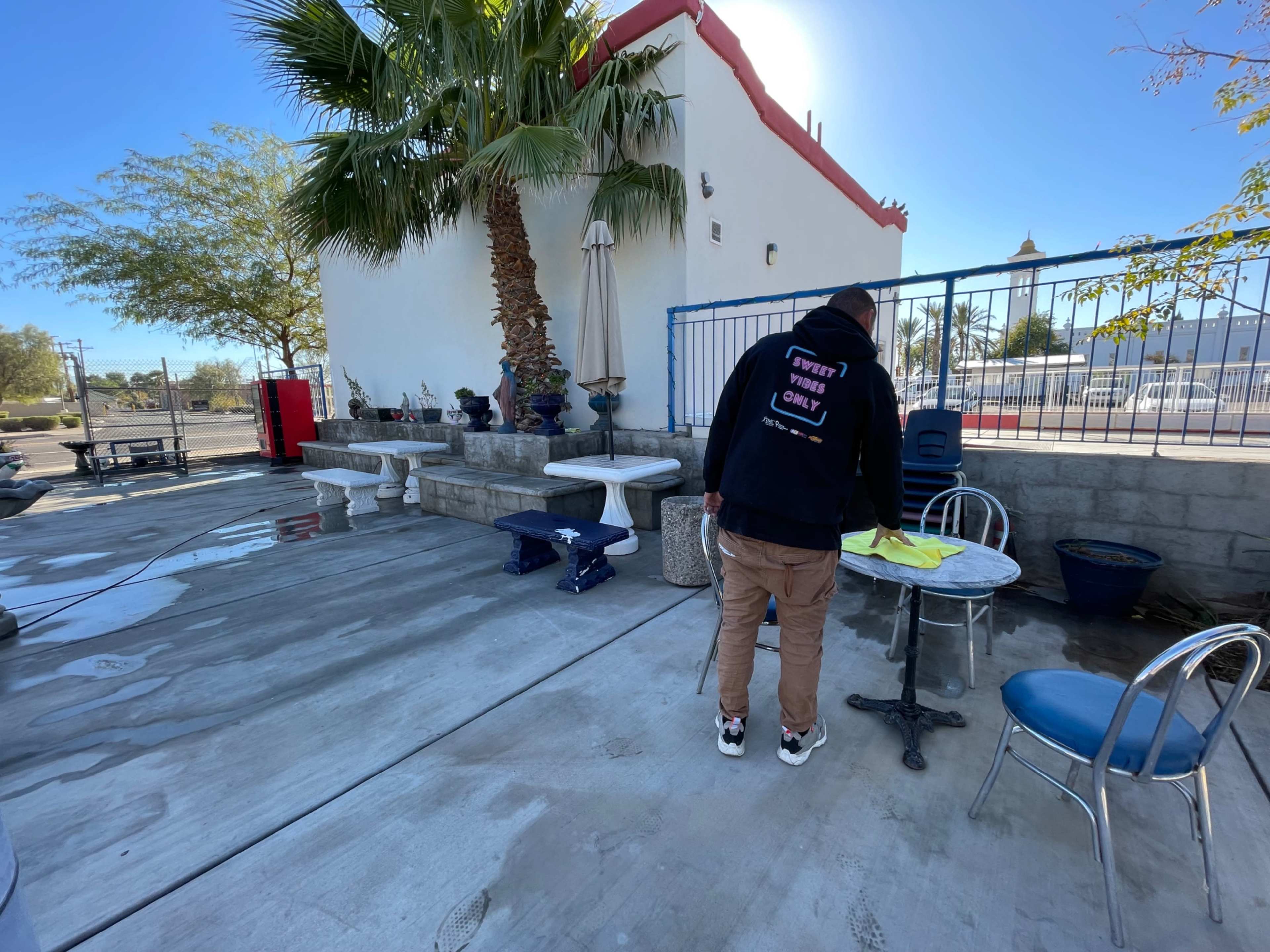 A person is wiping a table in a paved outdoor area surrounded by various plants and furniture.