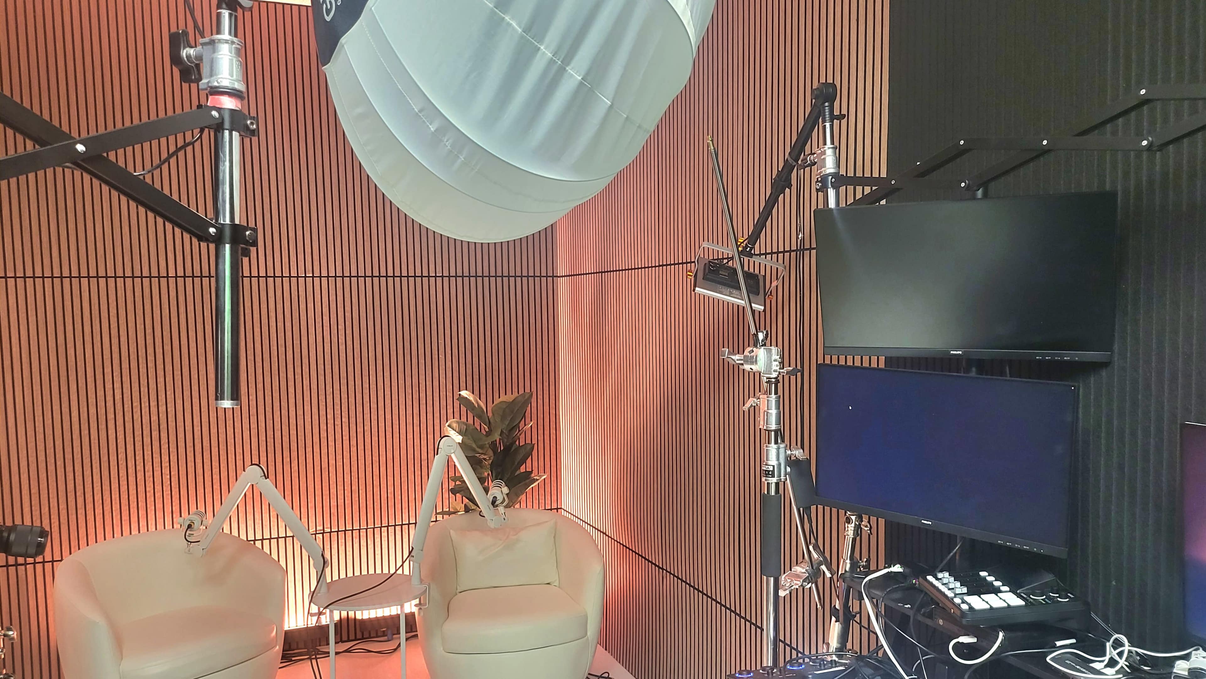 The image shows a modern recording or broadcasting studio with two white chairs, a large softbox light, multiple screens, and a vertical wooden panel wall.
