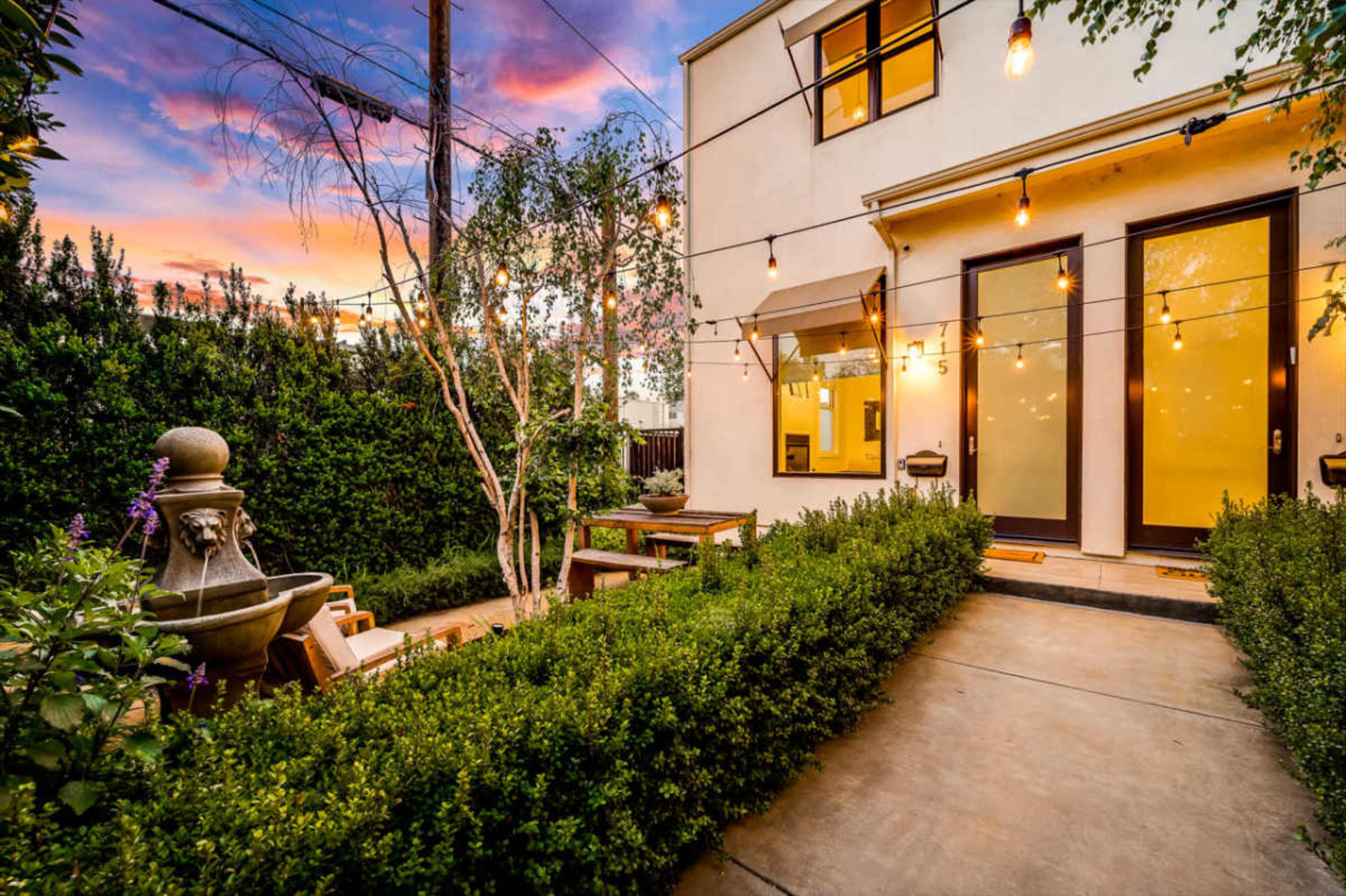 The image shows a well-lit backyard with neatly trimmed hedges, a central pathway, a stone fountain, and string lights hanging above outdoor seating near a house at sunset.