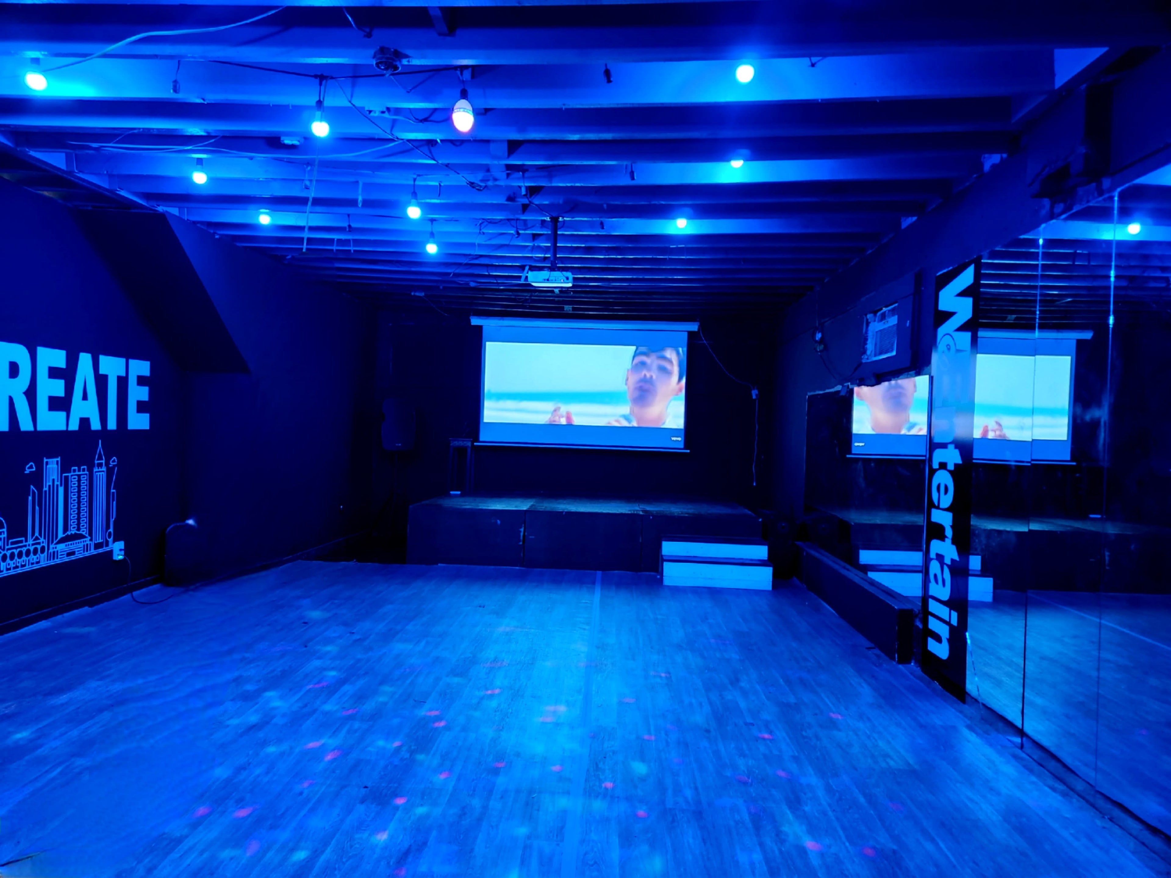 An empty room with blue lighting features a large screen displaying a video, a mirror on one wall, and the word "CREATE" painted on another wall.