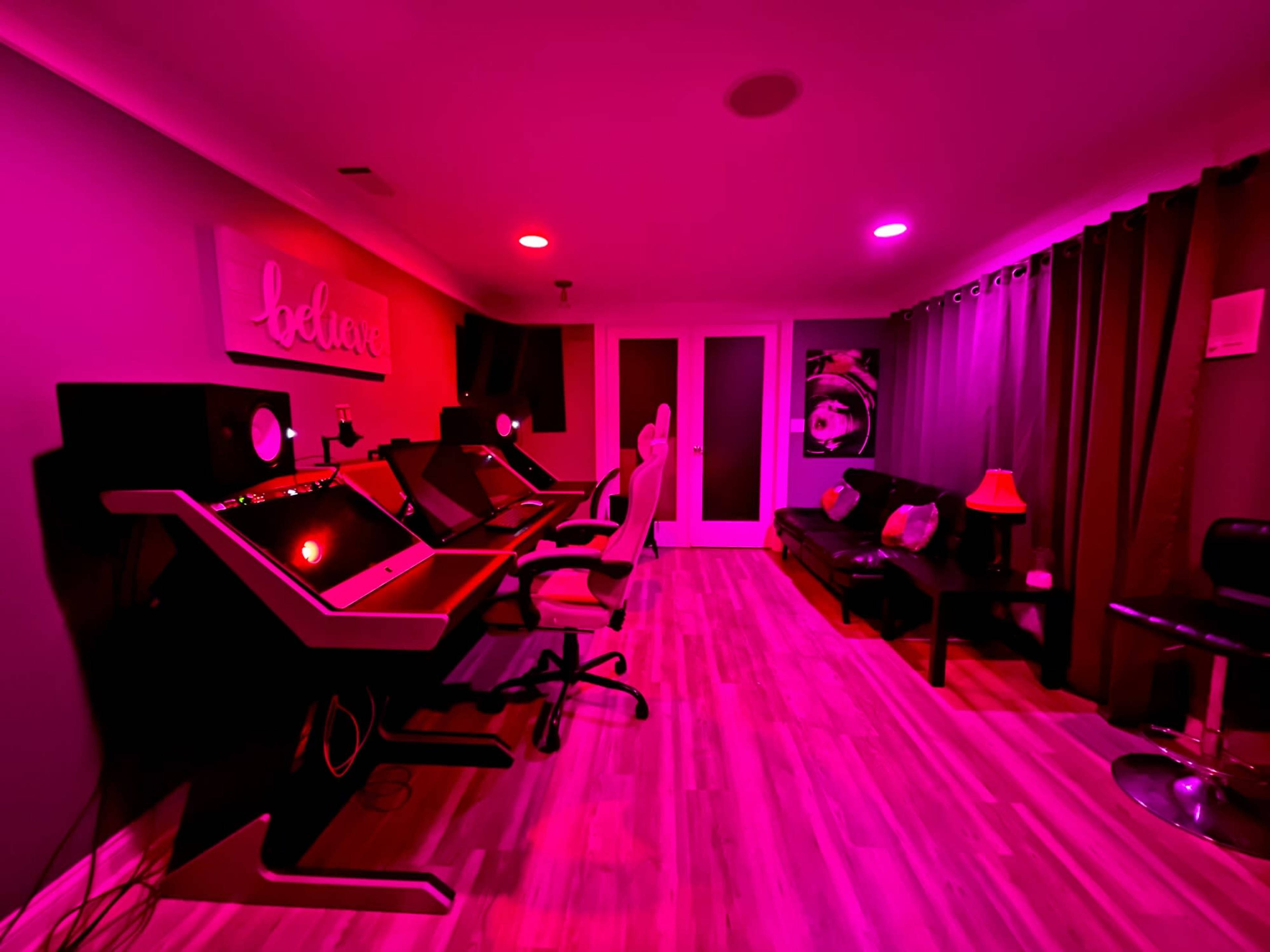 A music studio features two workstations with computer monitors in a room illuminated by pink and red lights, accompanied by a sofa and a chair.