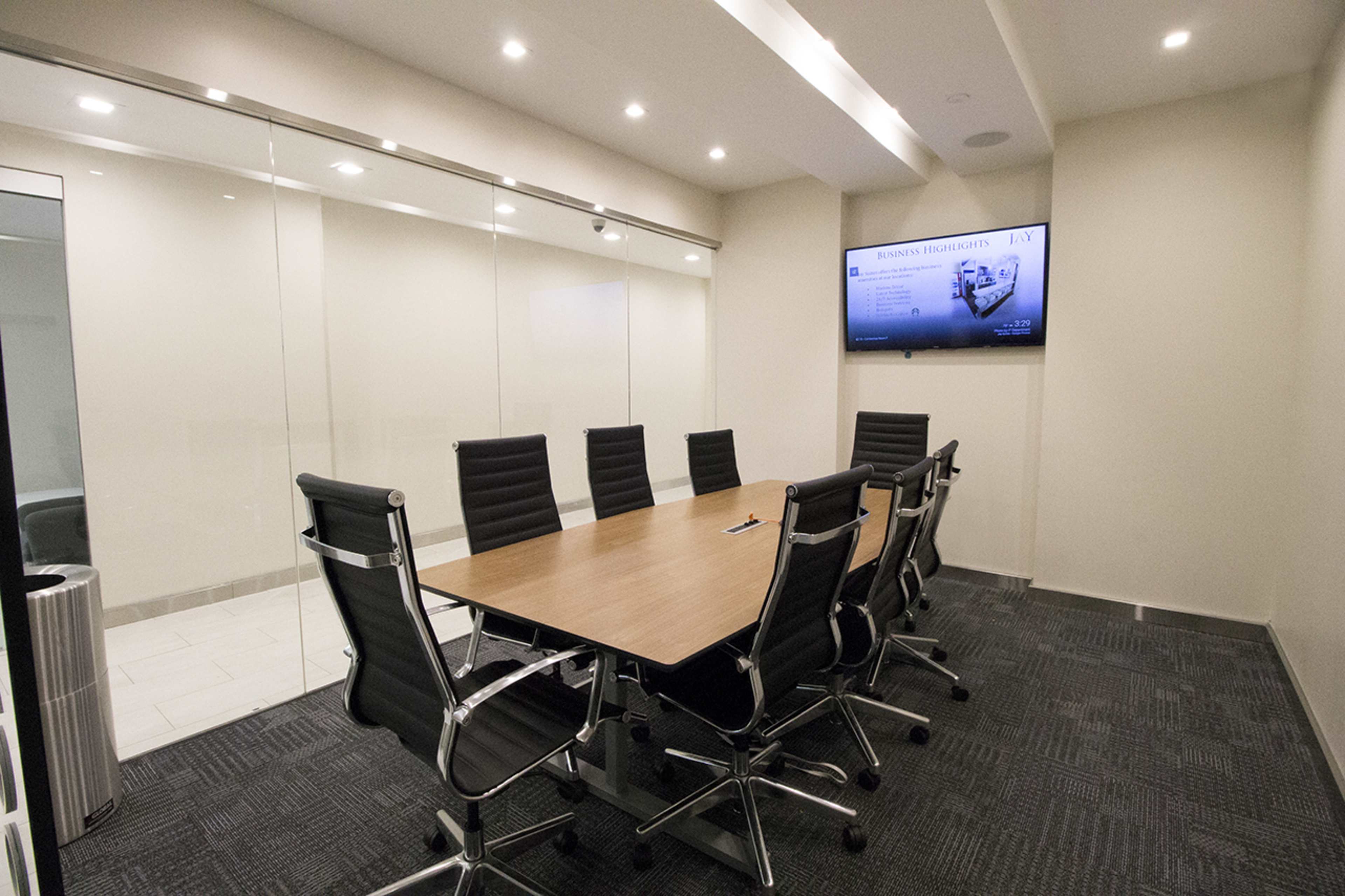 A conference room features a large wooden table surrounded by black leather office chairs, with a wall-mounted screen and glass walls.
