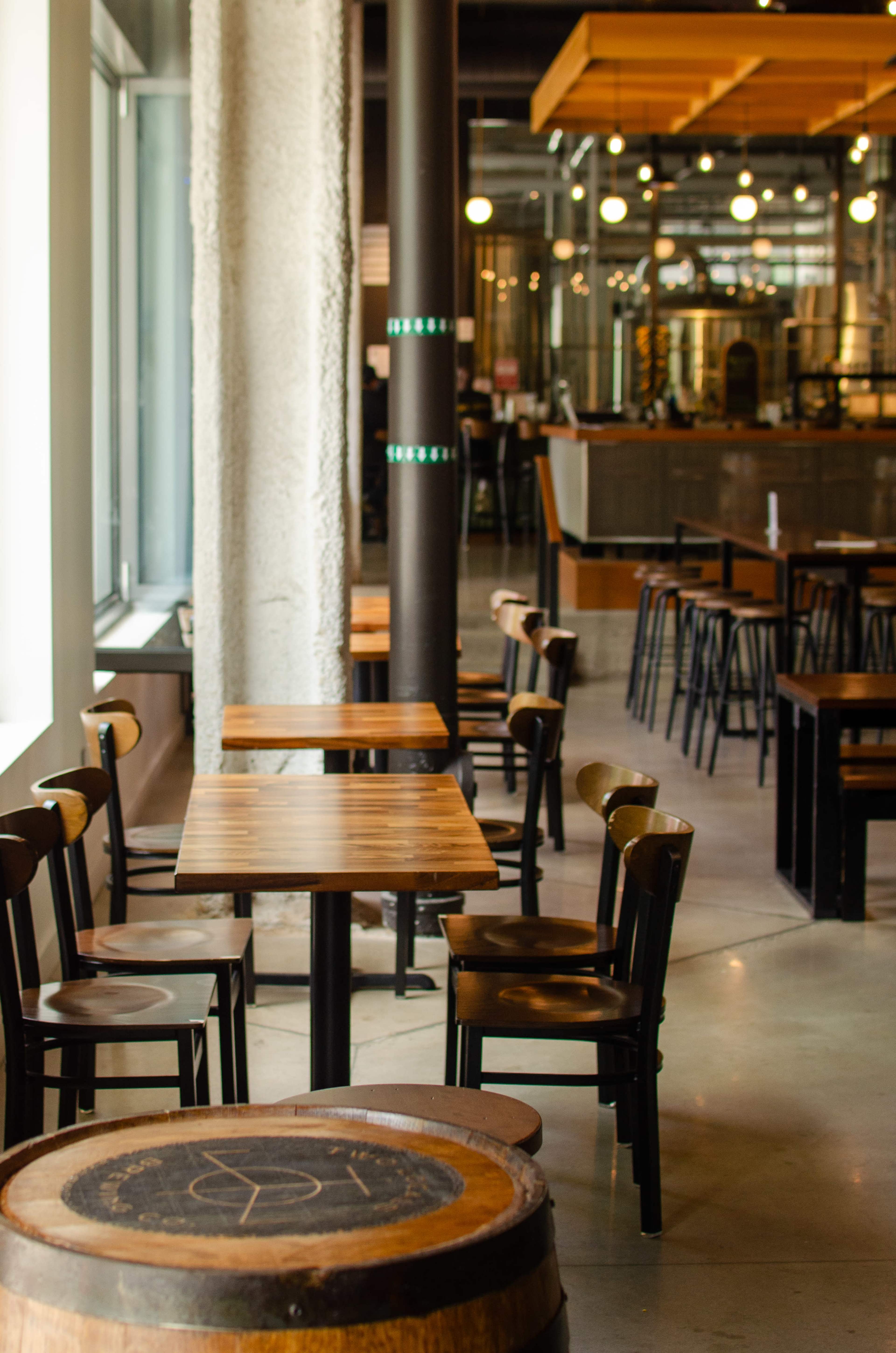 The image shows a modern café interior with wooden tables and chairs arranged around a central area, featuring large windows and a bar in the background.