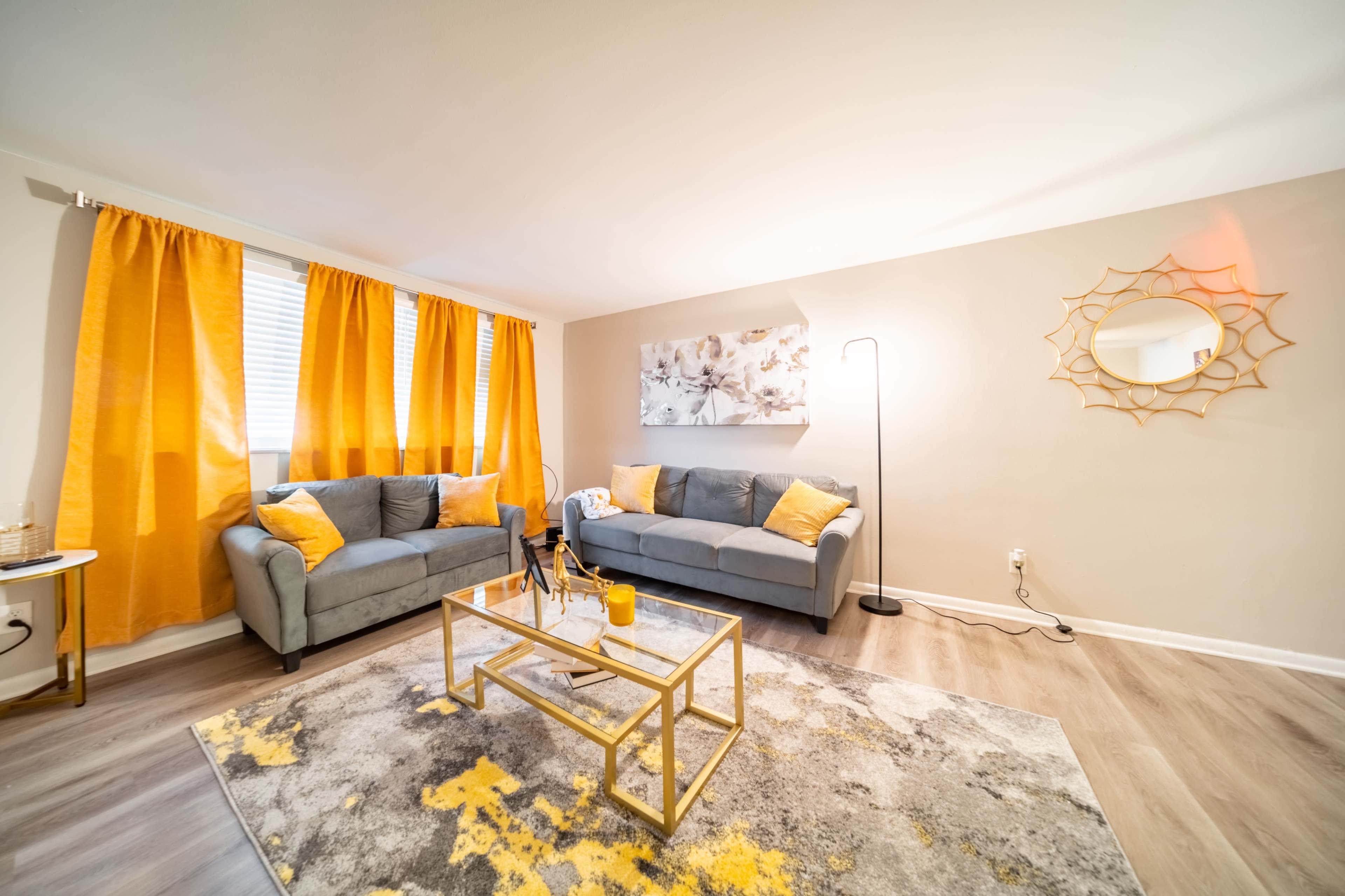 The living room features two gray sofas with yellow pillows, yellow curtains, a decorative gold mirror, and a patterned rug under a glass coffee table.