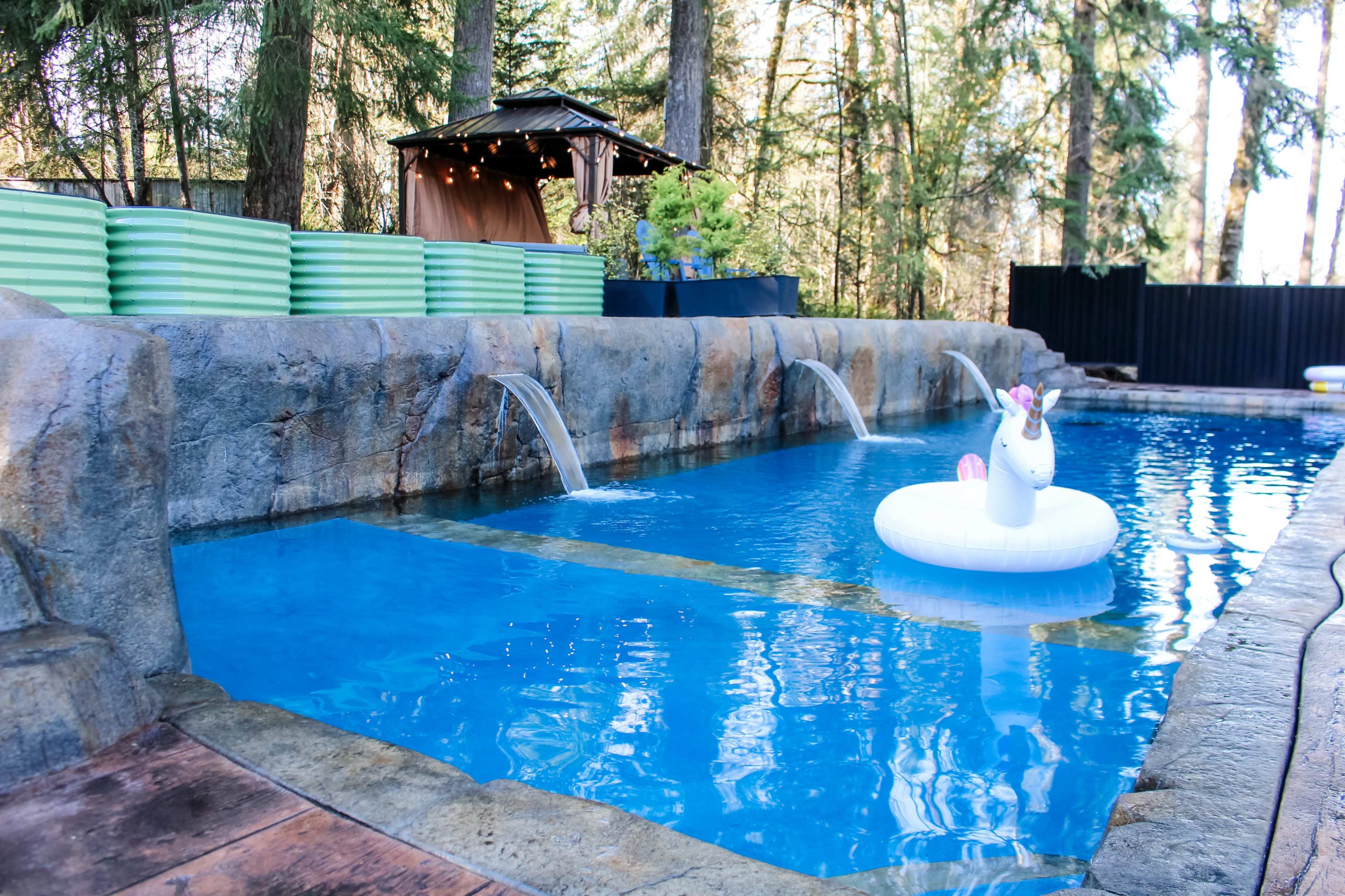 The image shows a swimming pool surrounded by large stone features, a unicorn float in the water, and greenery in the background.