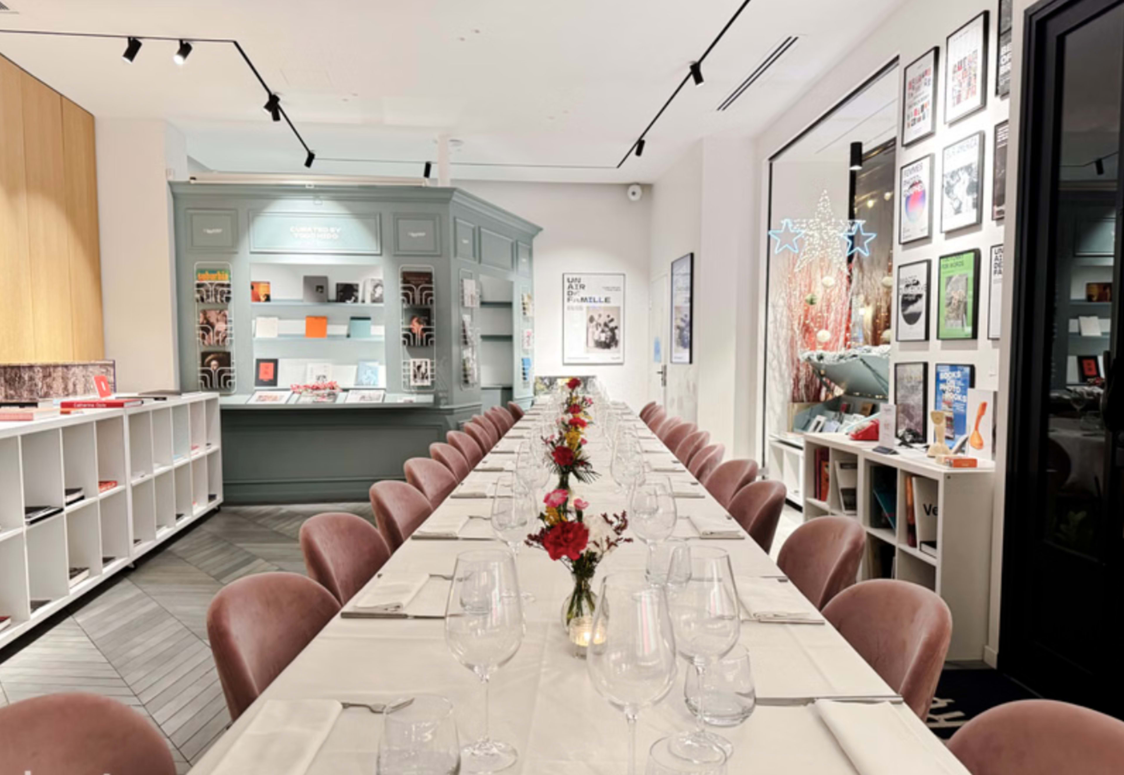 The image shows a modern dining setup with a long table adorned with pink chairs, neatly arranged white tablecloths, glasses, and small flower arrangements, all in a well-lit room featuring shelves filled with books and decorative items.