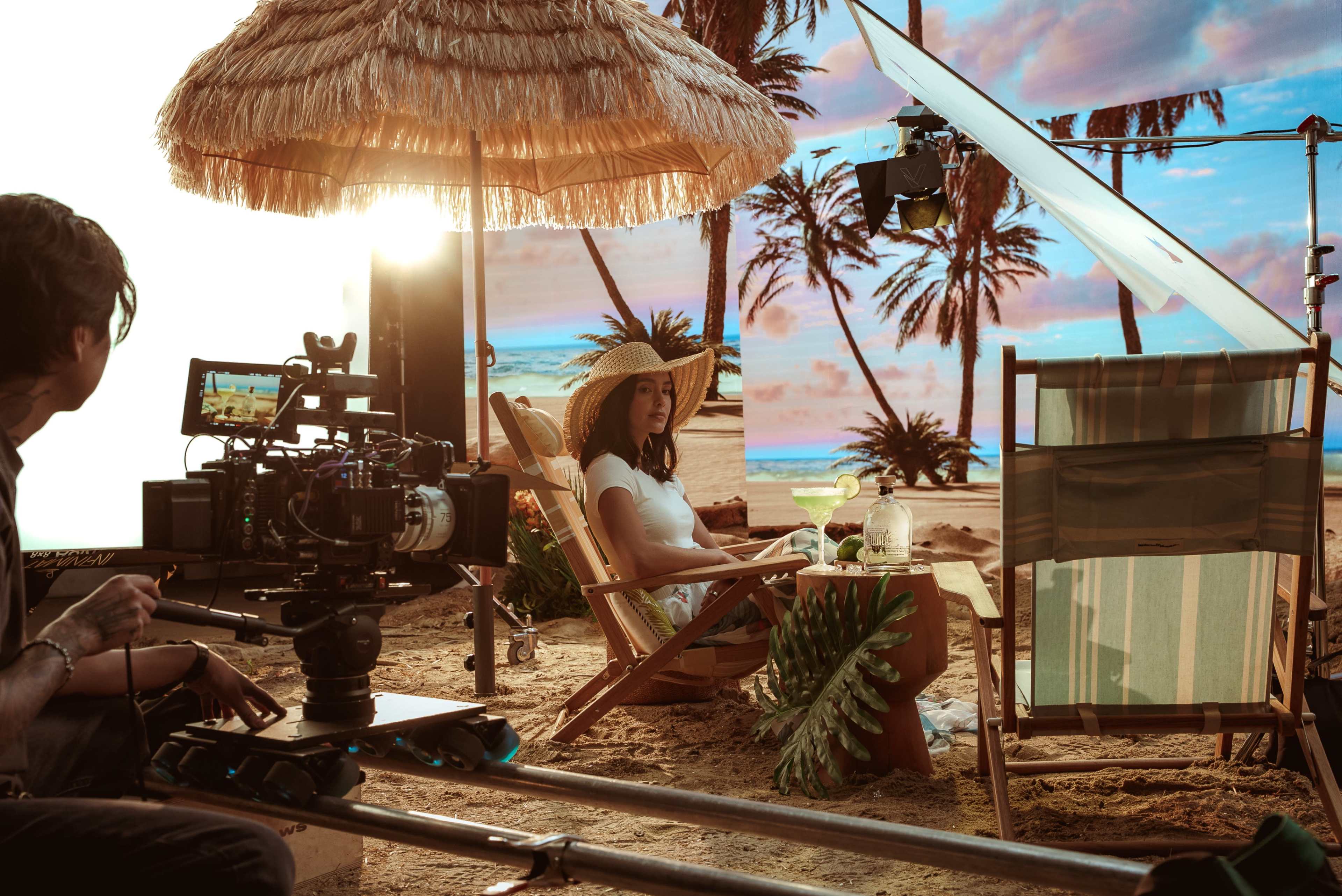 A film crew sets up a beach scene with a woman seated under a straw umbrella, surrounded by palm trees and a backdrop of colorful clouds.