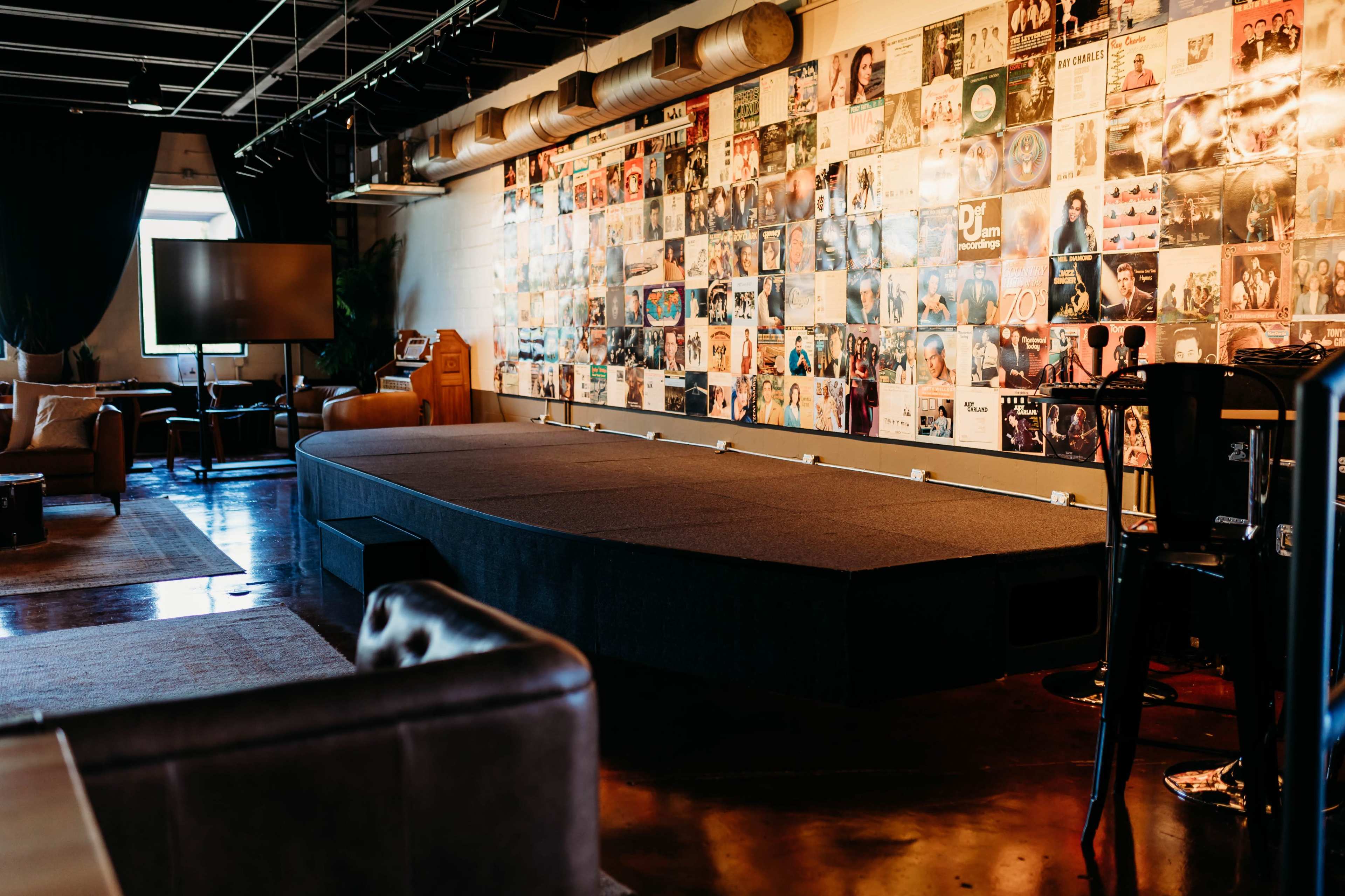 The image shows a stage with a dark carpet in a room decorated with a collage of vintage record album covers on the wall.