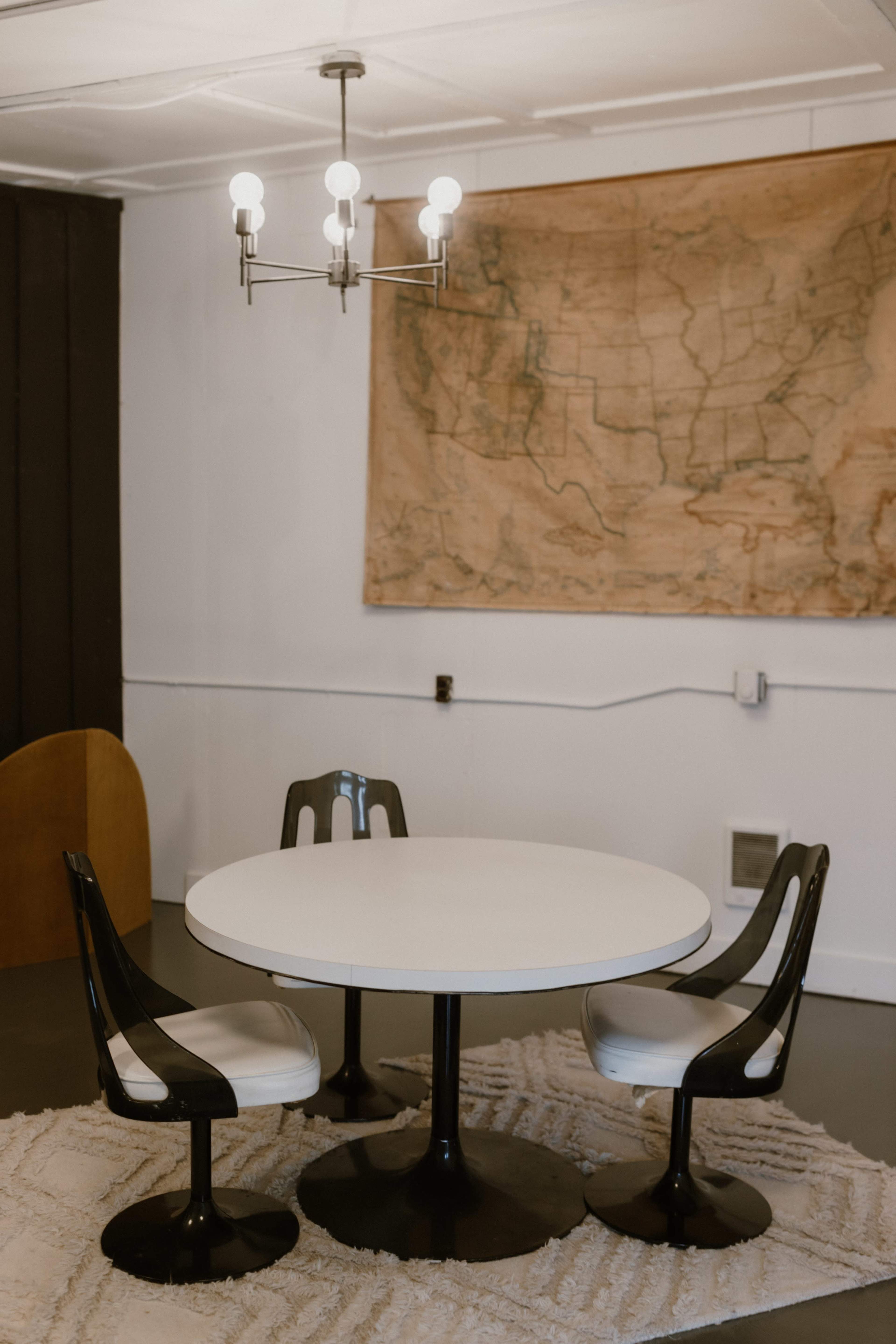 A round white table is surrounded by three black and white chairs in a room featuring a large map on the wall.