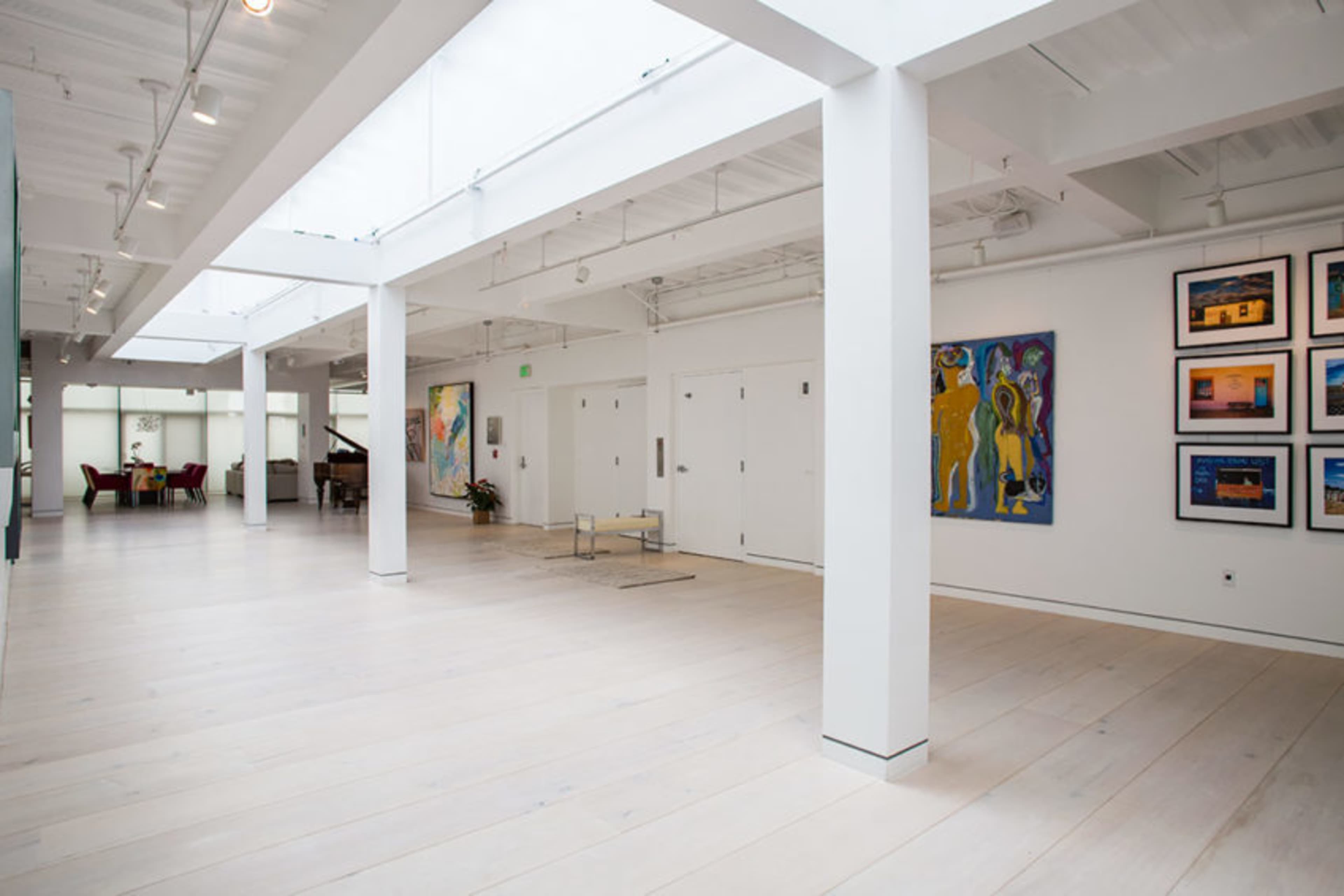 A spacious, well-lit gallery with white walls, wooden floors, and paintings displayed on the walls.