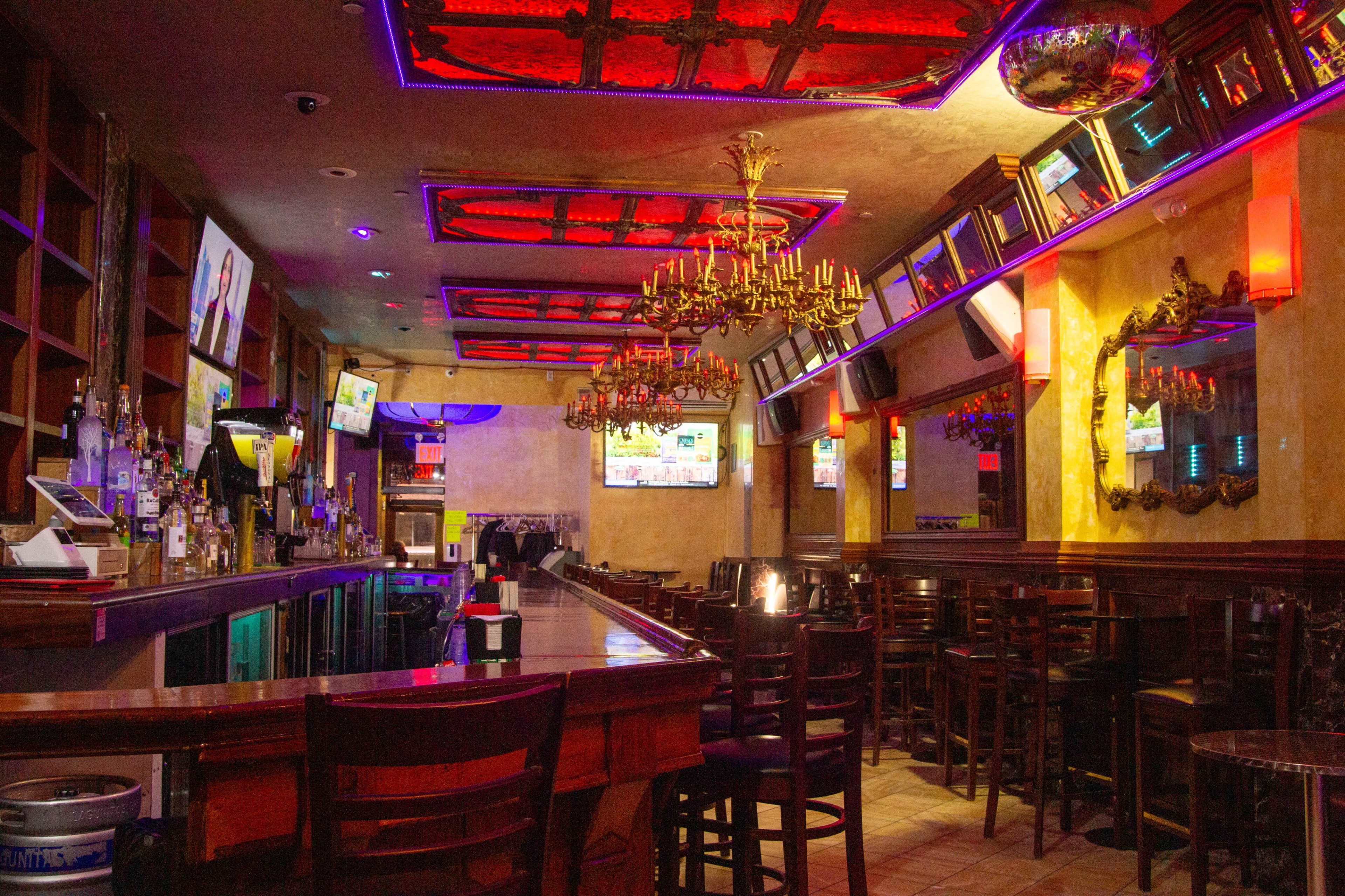 The interior of a bar features a long wooden counter, high chairs, chandeliers, and several TV screens on the walls, all illuminated by colorful lights.