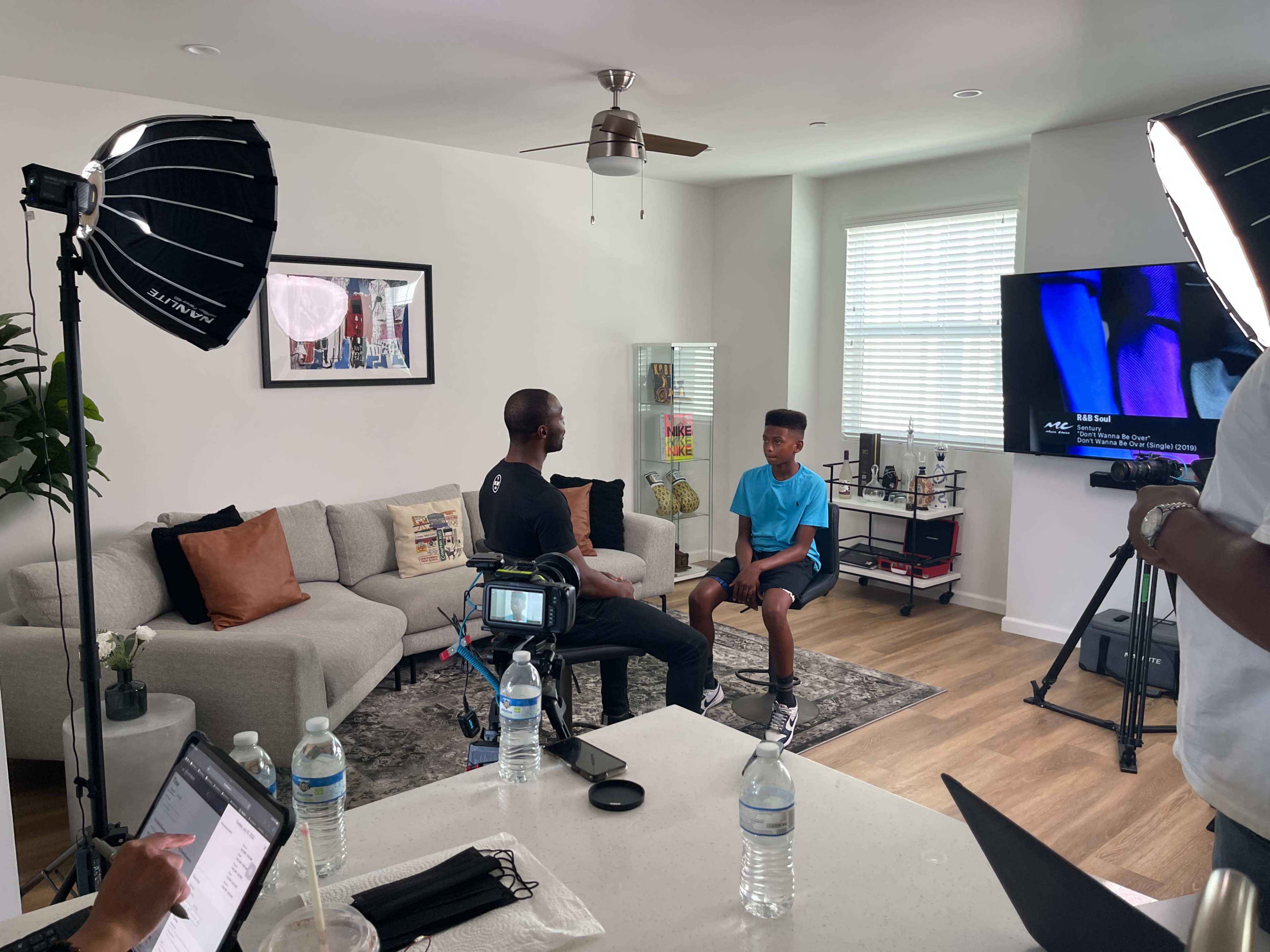 A man and a boy are seated across from each other in a brightly lit living room during an interview setup, with cameras and lighting equipment nearby.