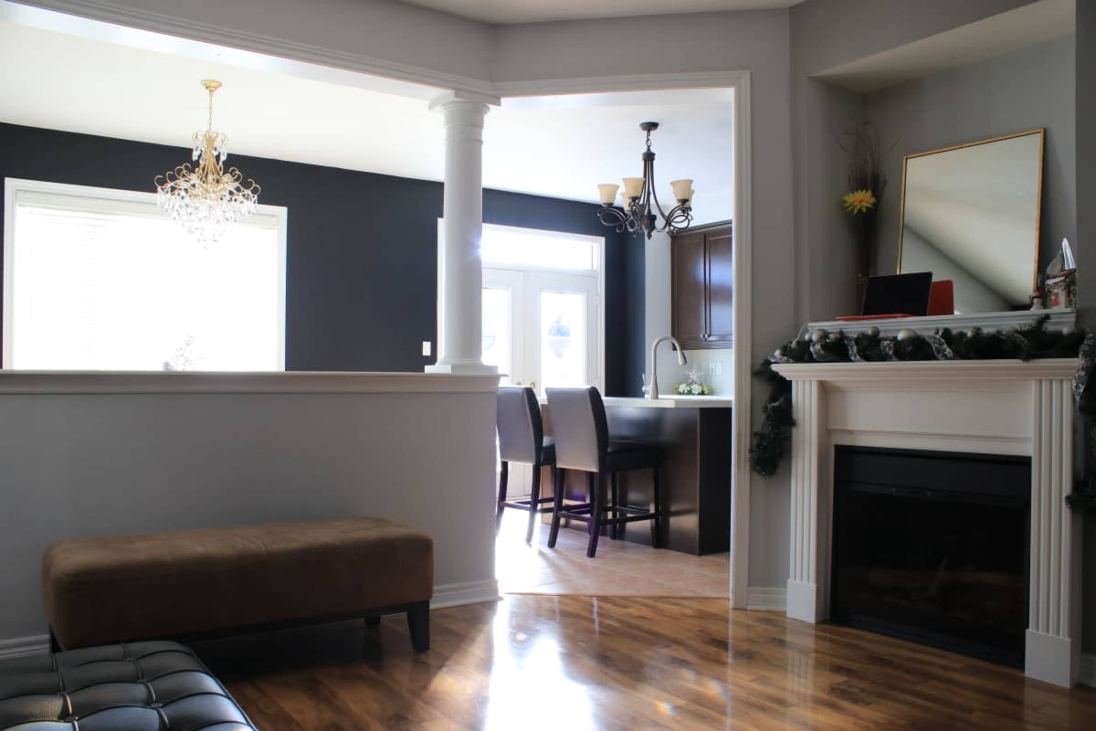 The image shows a living area with a brown ottoman, a fireplace, and an open view into a dining room featuring dark walls and a chandelier.