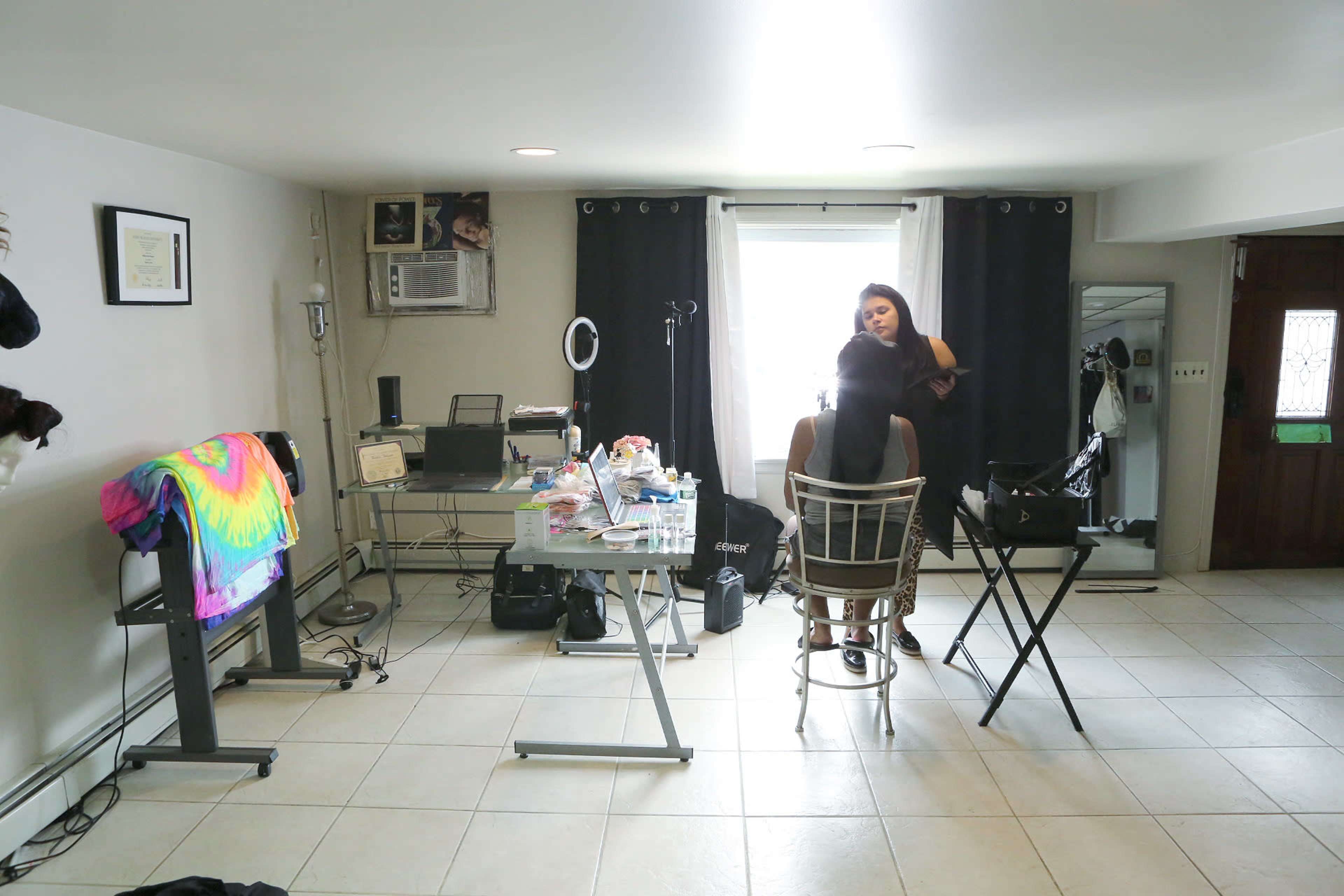 A makeup artist applies makeup to a seated client in a well-lit room with various beauty equipment and a mirror.
