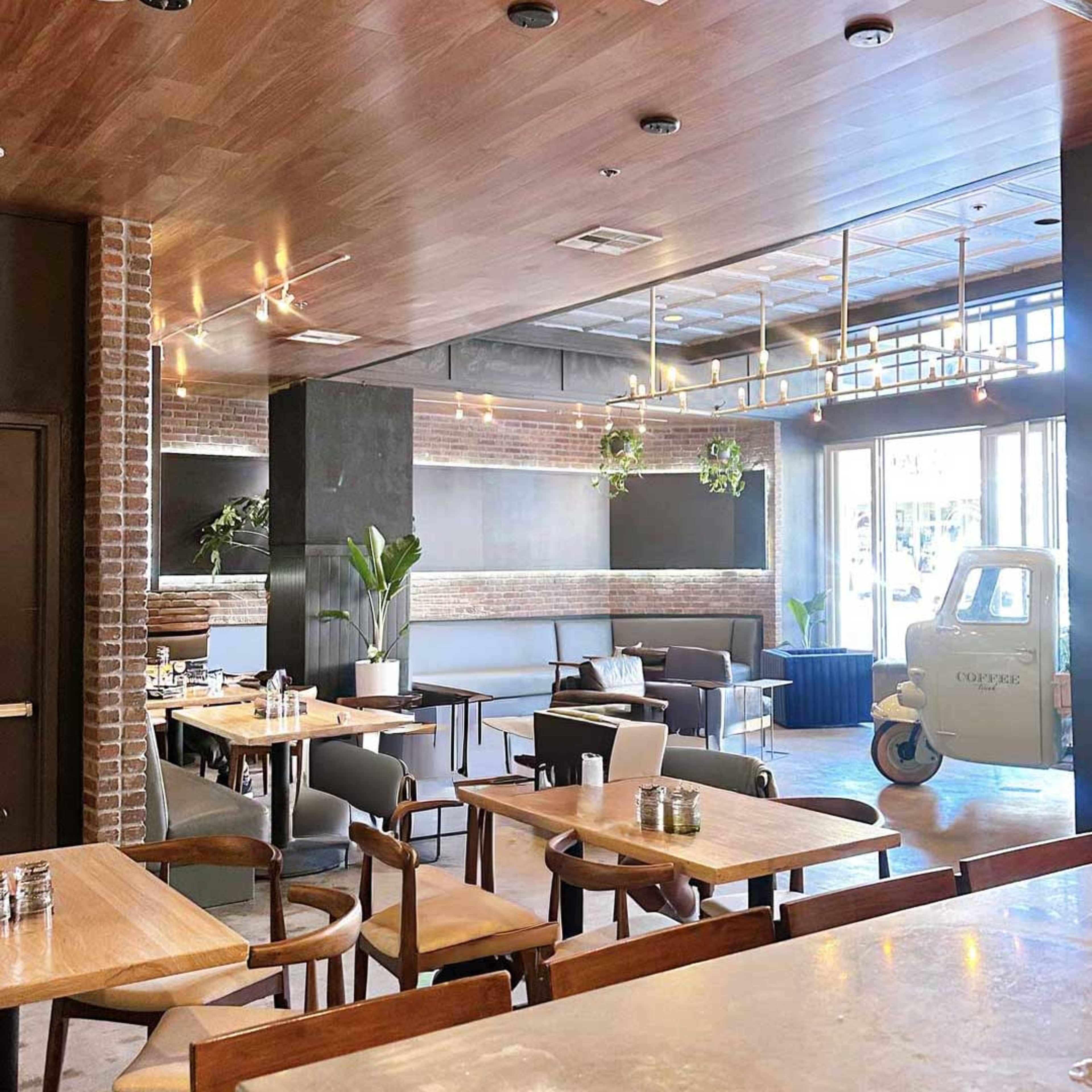 The image shows a modern restaurant interior with wooden tables, minimalistic décor, and large windows allowing natural light.