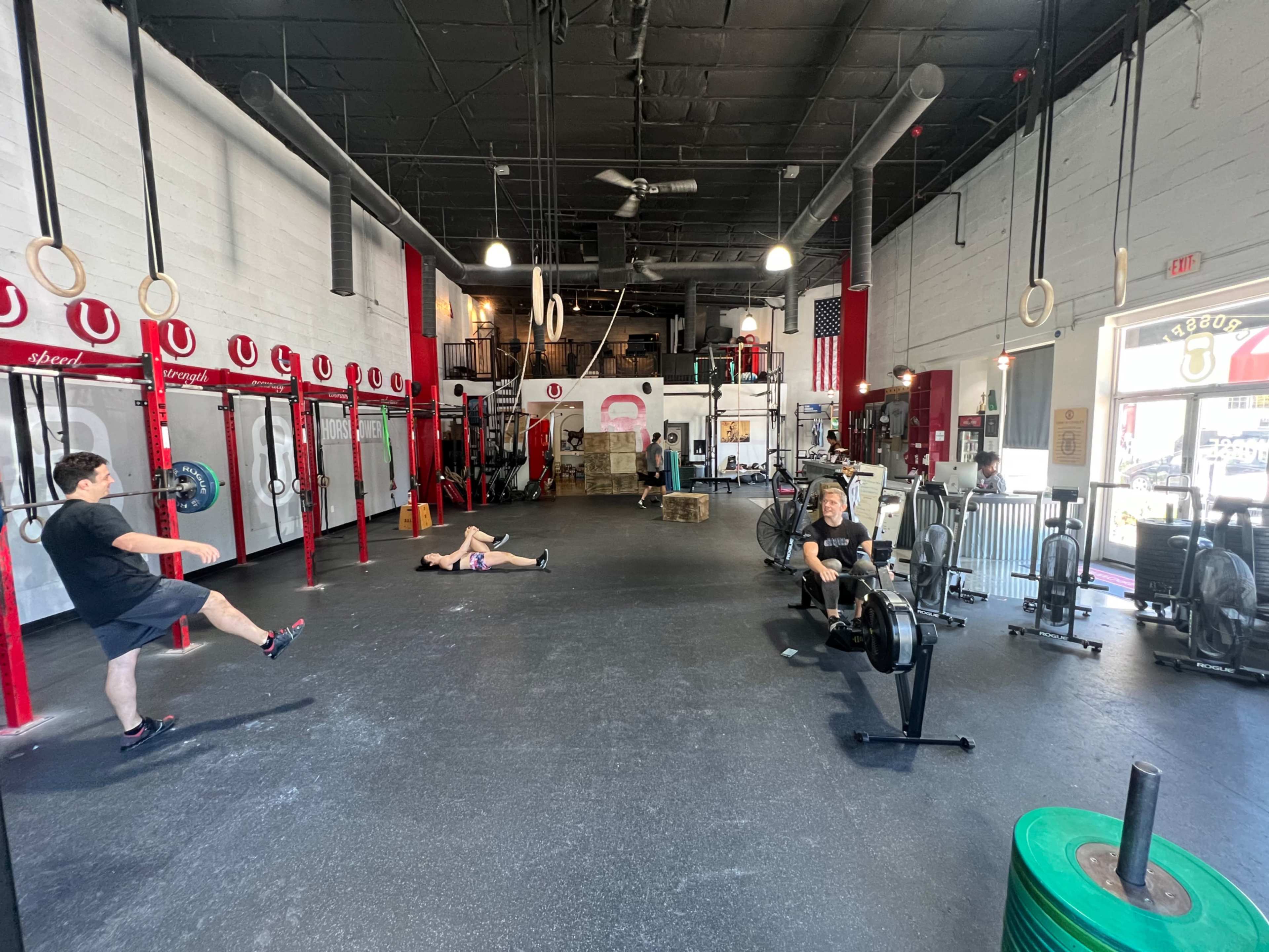 A spacious gym interior features individuals exercising with rowing machines and rings, while one person lies on the floor.
