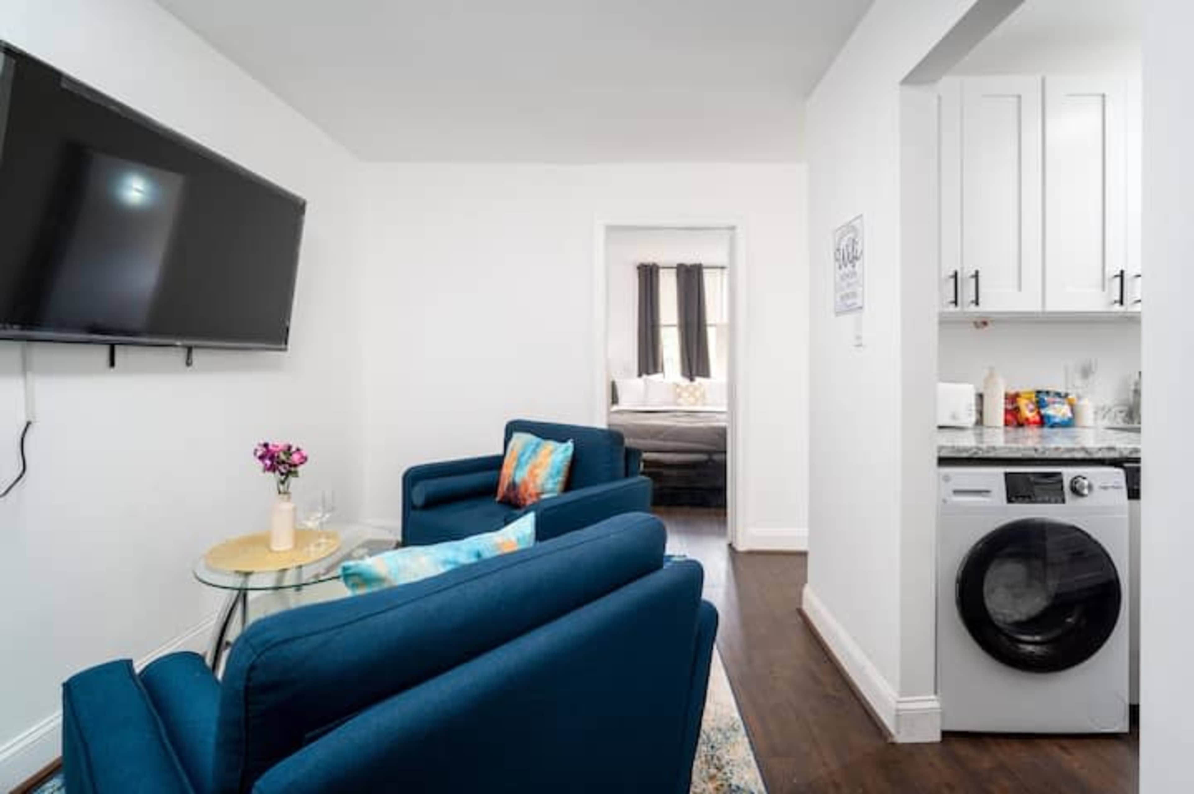 A small living area featuring two blue chairs, a round glass table, a wall-mounted TV, and a doorway leading to a bedroom and a kitchenette with a washing machine.