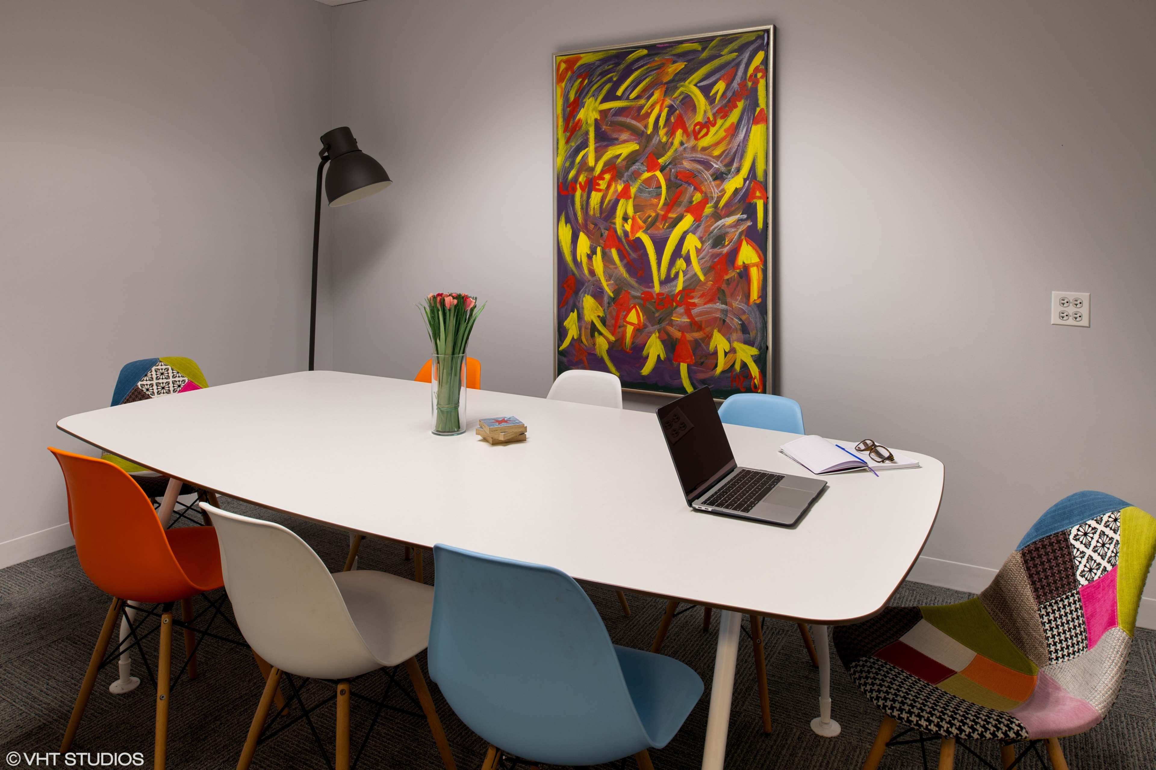 A modern conference room features a long white table surrounded by multicolored chairs, with a laptop and notebook on the table, and a large abstract painting on the wall.