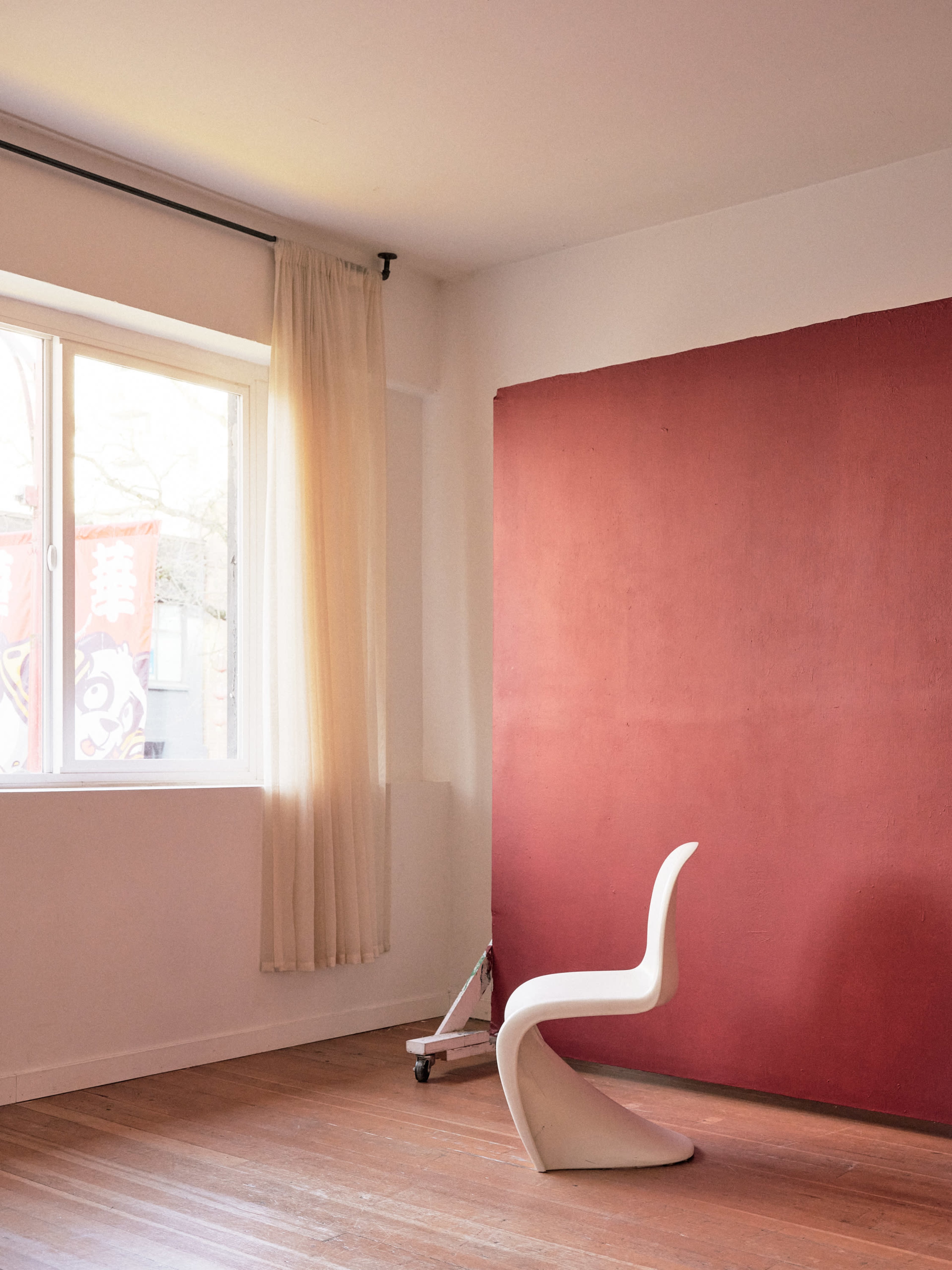 A white chair is positioned next to a window in a room with a red accent wall and light wooden flooring.