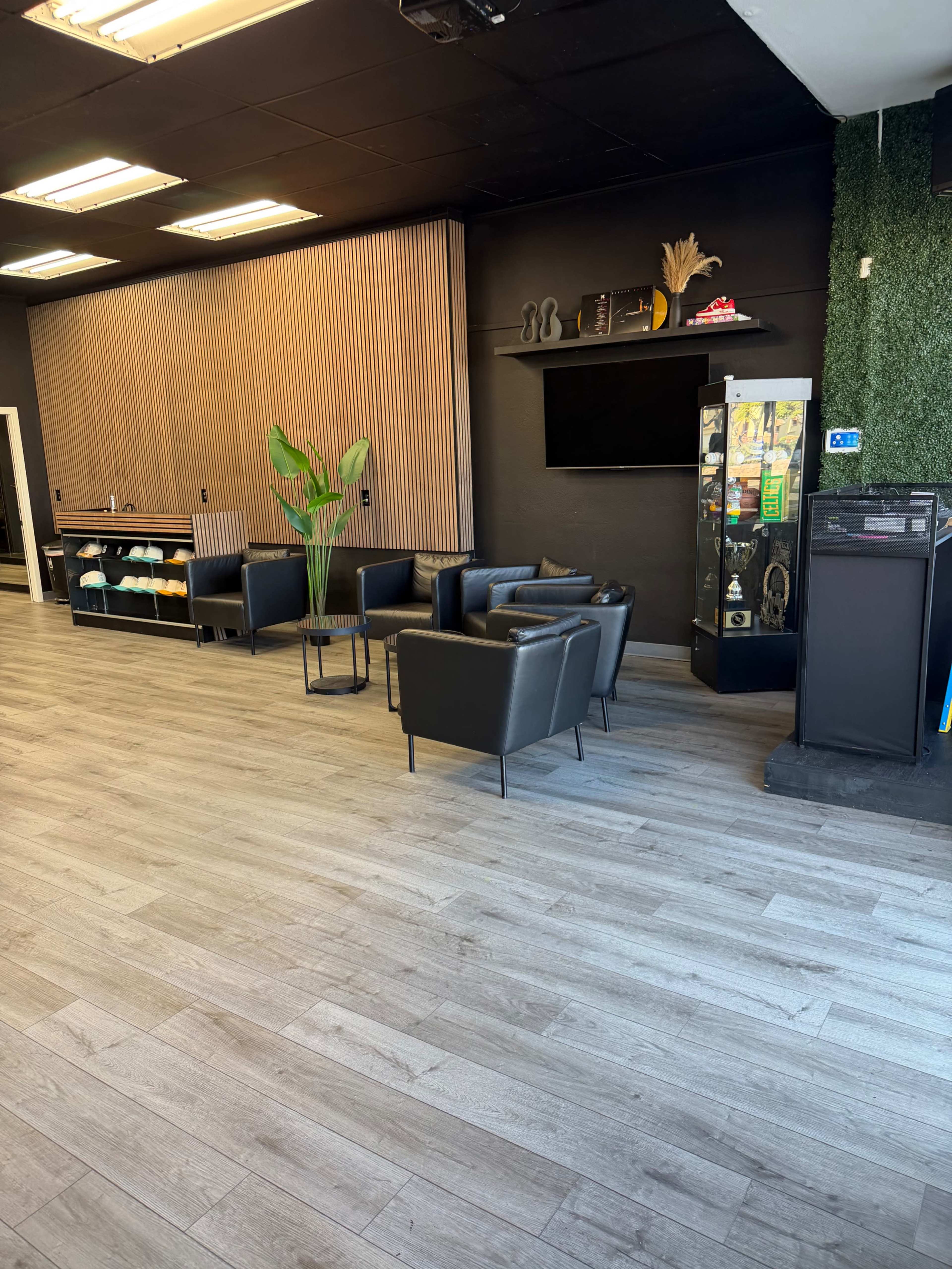 The image shows a modern waiting area with black chairs, a small table, and a green plant, set against a backdrop of wooden paneling and a television screen.
