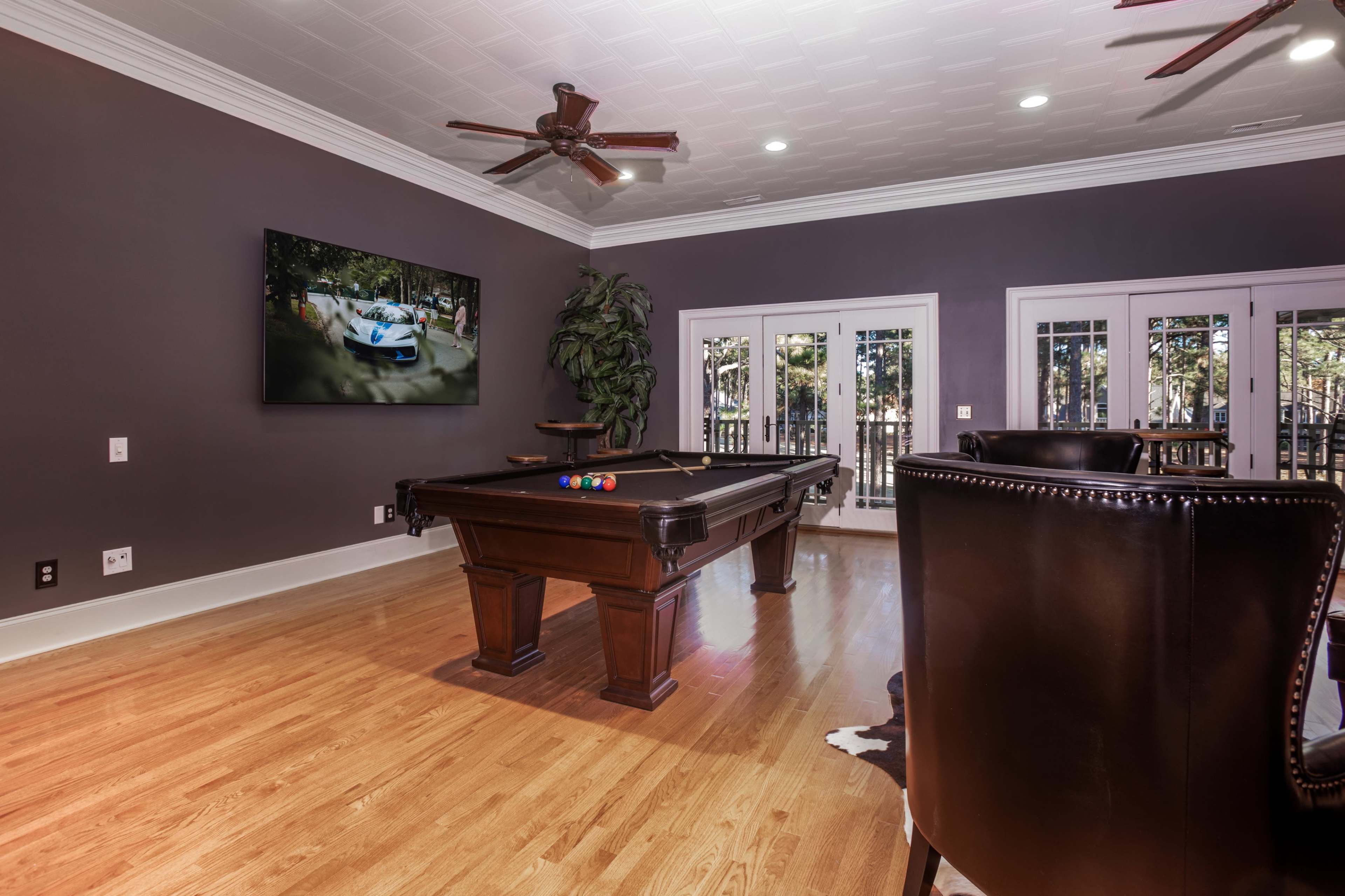 A room with a pool table centered beneath a television screen, surrounded by windows and a plush seating area.