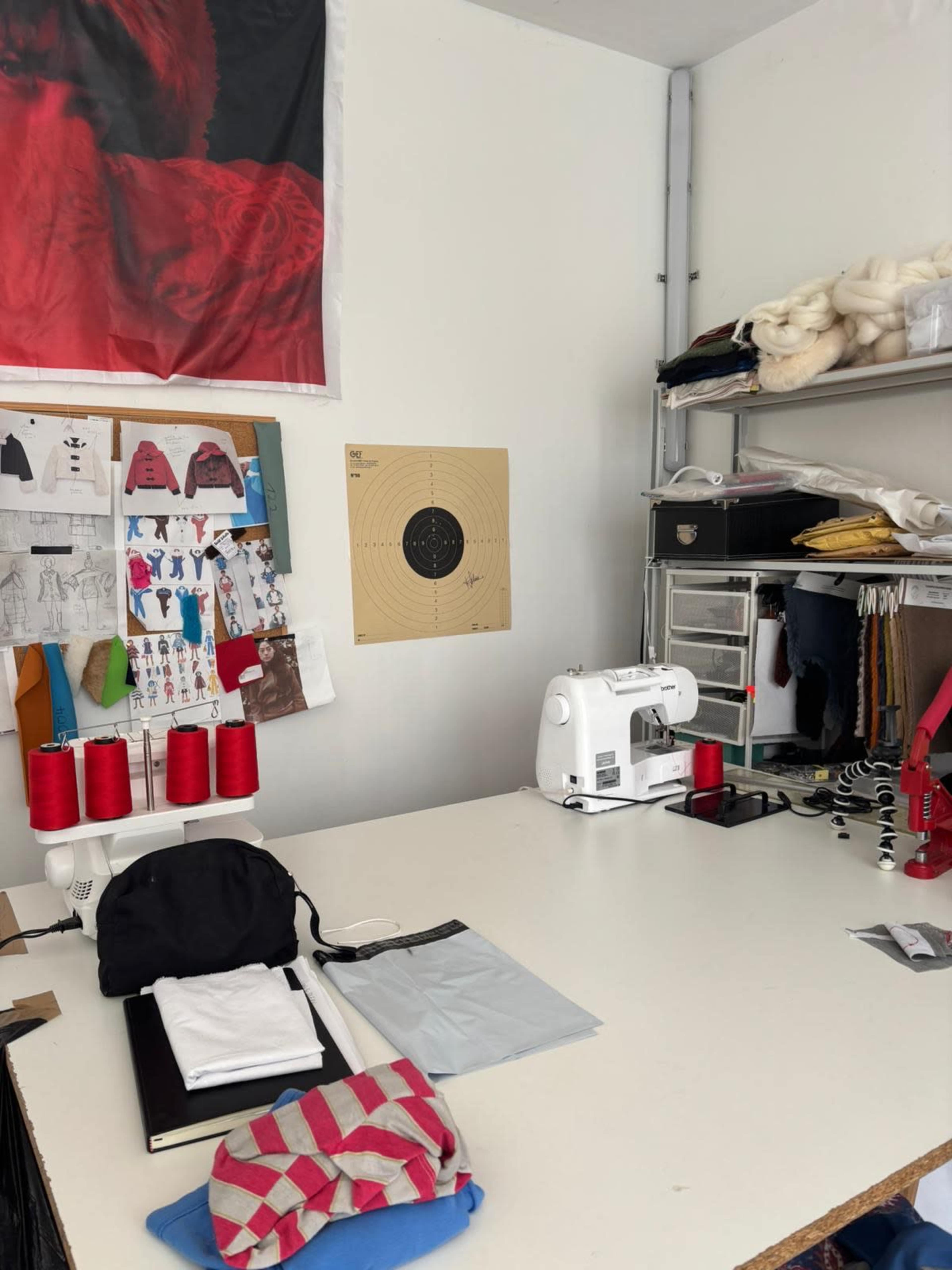 The image shows a sewing workspace featuring a white sewing machine, spools of red thread, sketches on the wall, and various fabric items on a table.