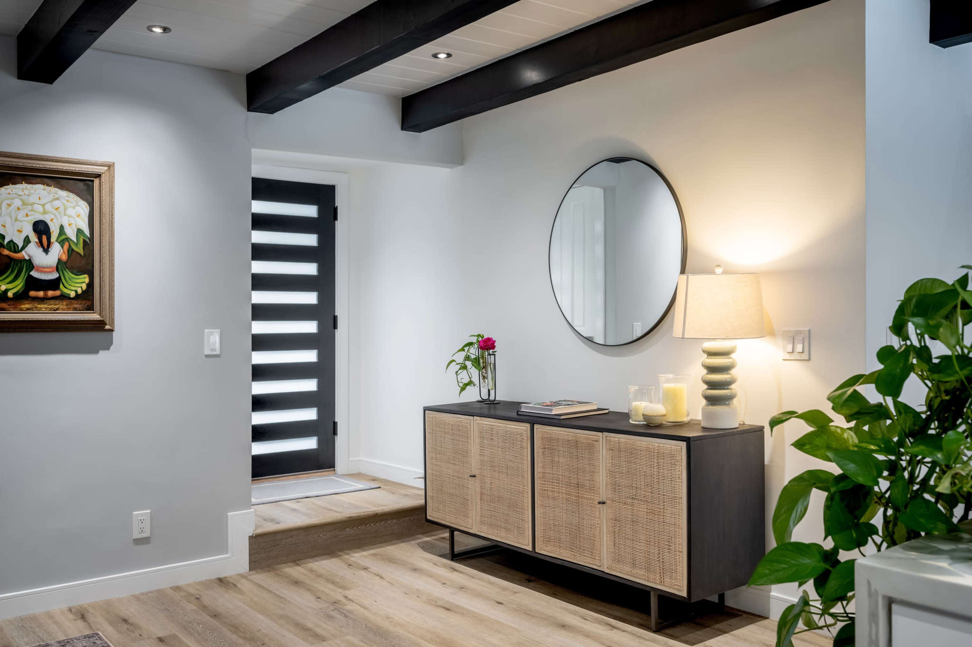 The image shows a modern entryway featuring a console table with a lamp, a round mirror, a piece of artwork on the wall, and a door with horizontal slats.