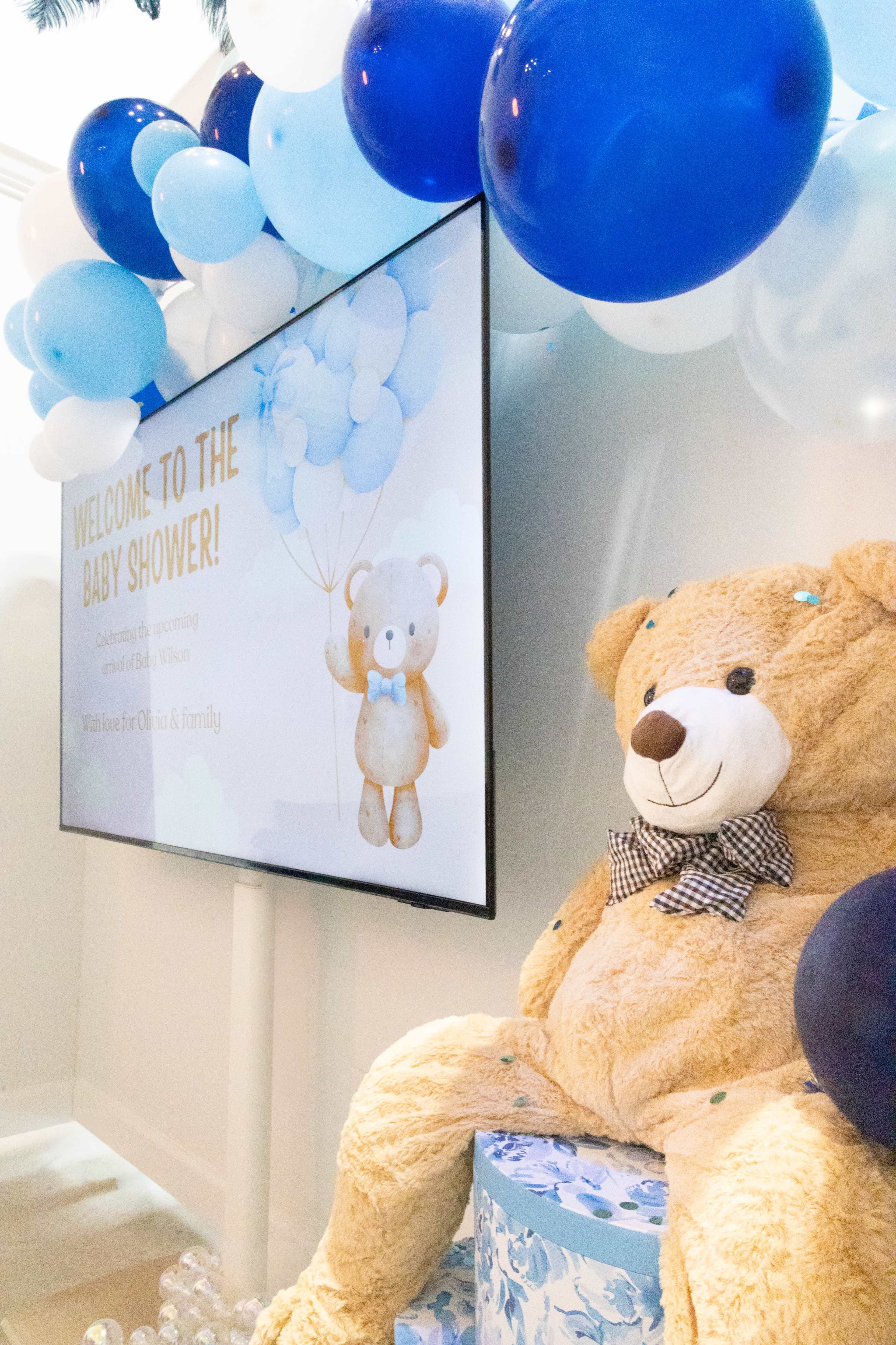 A large screen displays a welcome message for a baby shower, accompanied by a stuffed bear and blue balloon decorations.