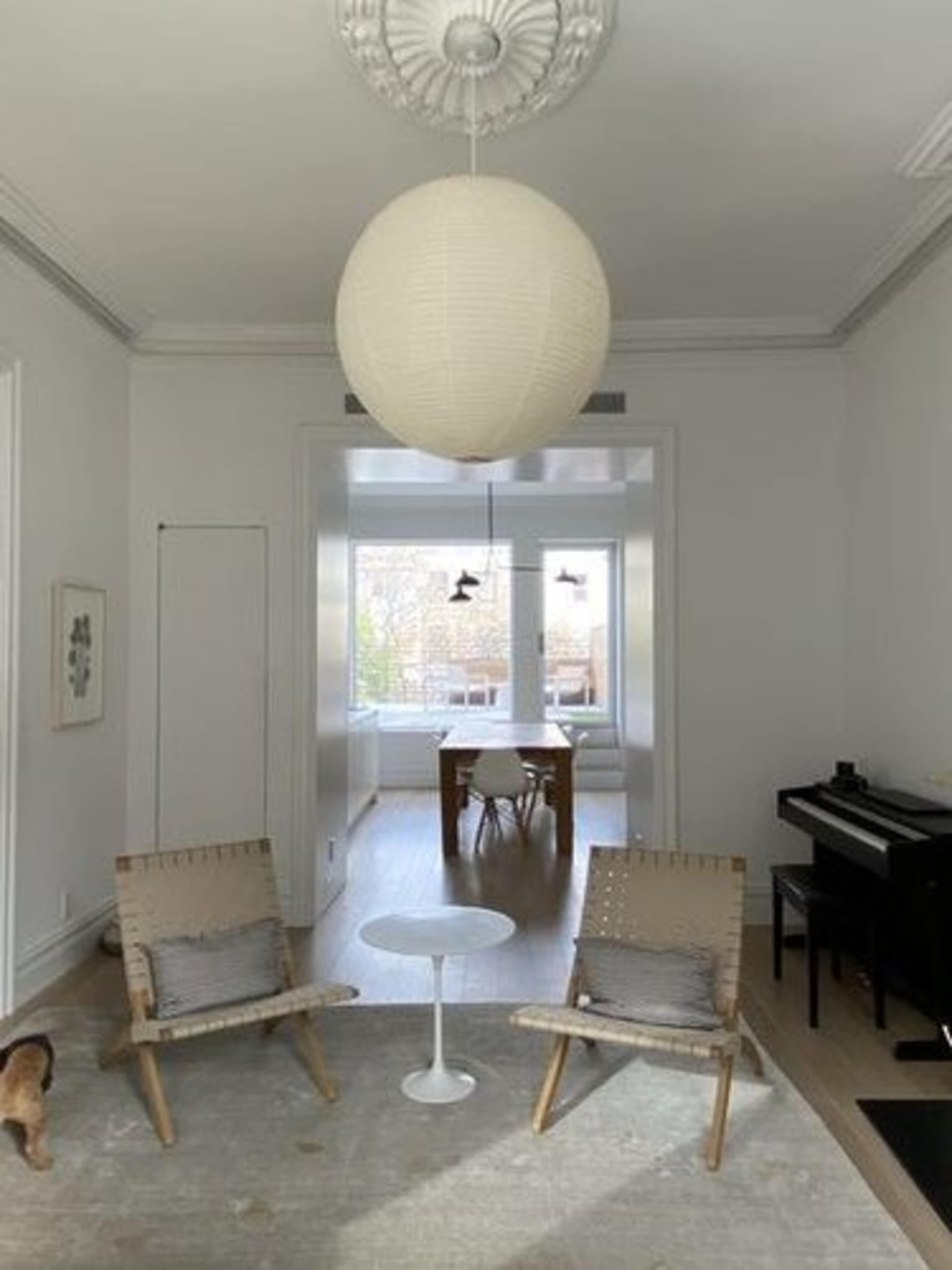 A spacious, modern living area with two woven chairs, a round side table, and a grand piano, leading into a bright dining space with a large window.