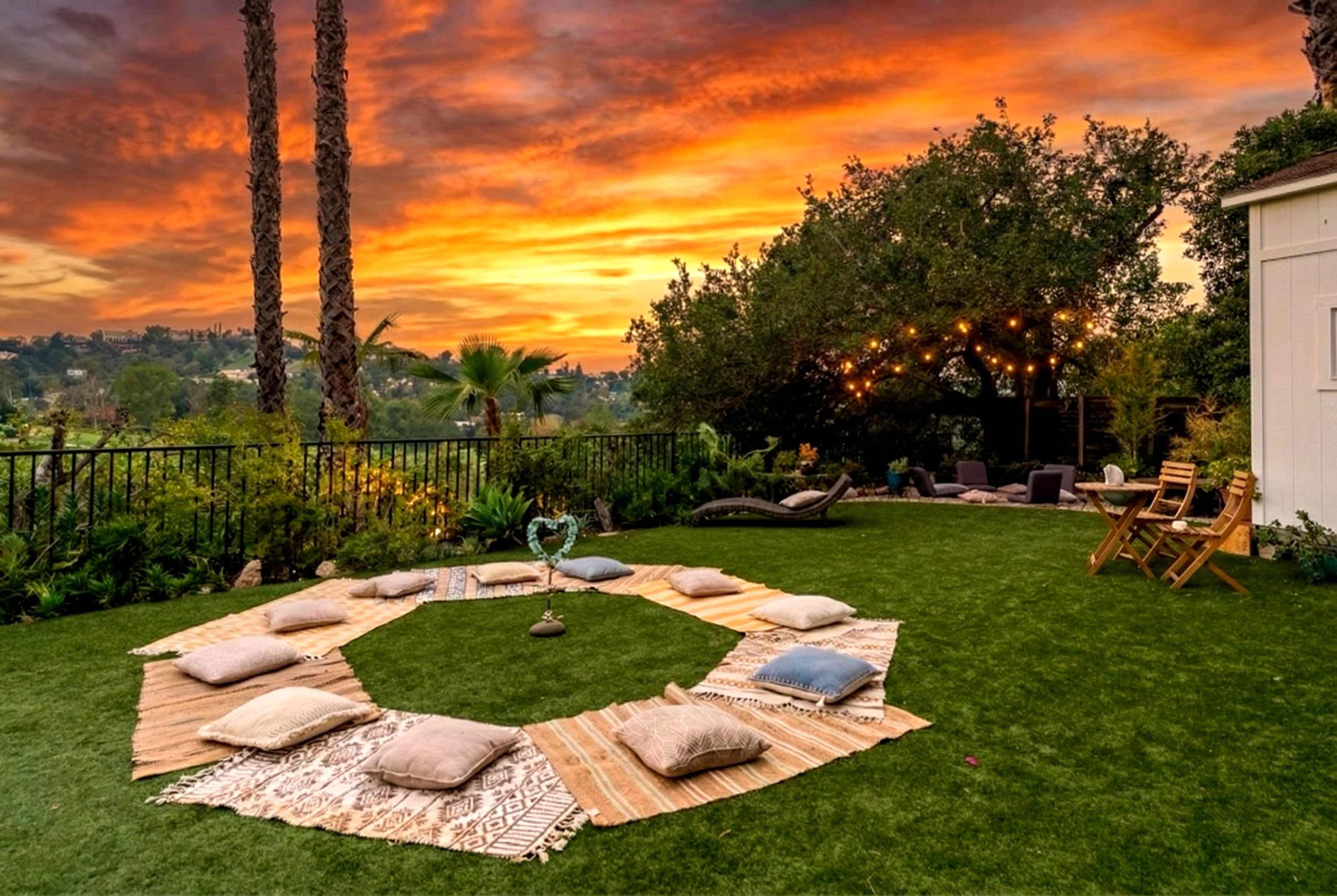 The image shows a circular arrangement of cushions and rugs on green grass, set against a colorful sunset and palm trees in the background.