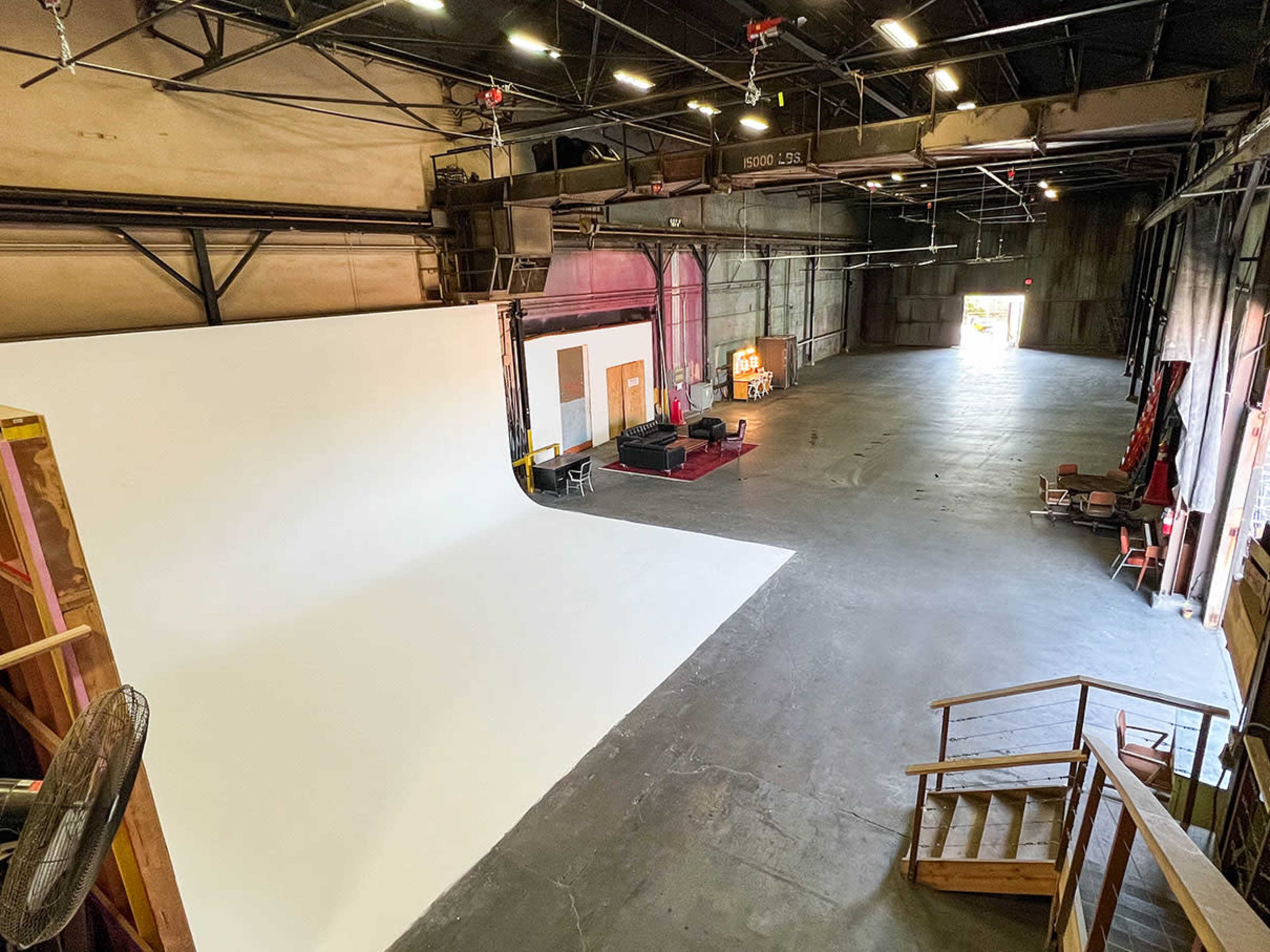 The image shows a large, empty studio space with a white backdrop, various furniture, and an open area illuminated by overhead lights.