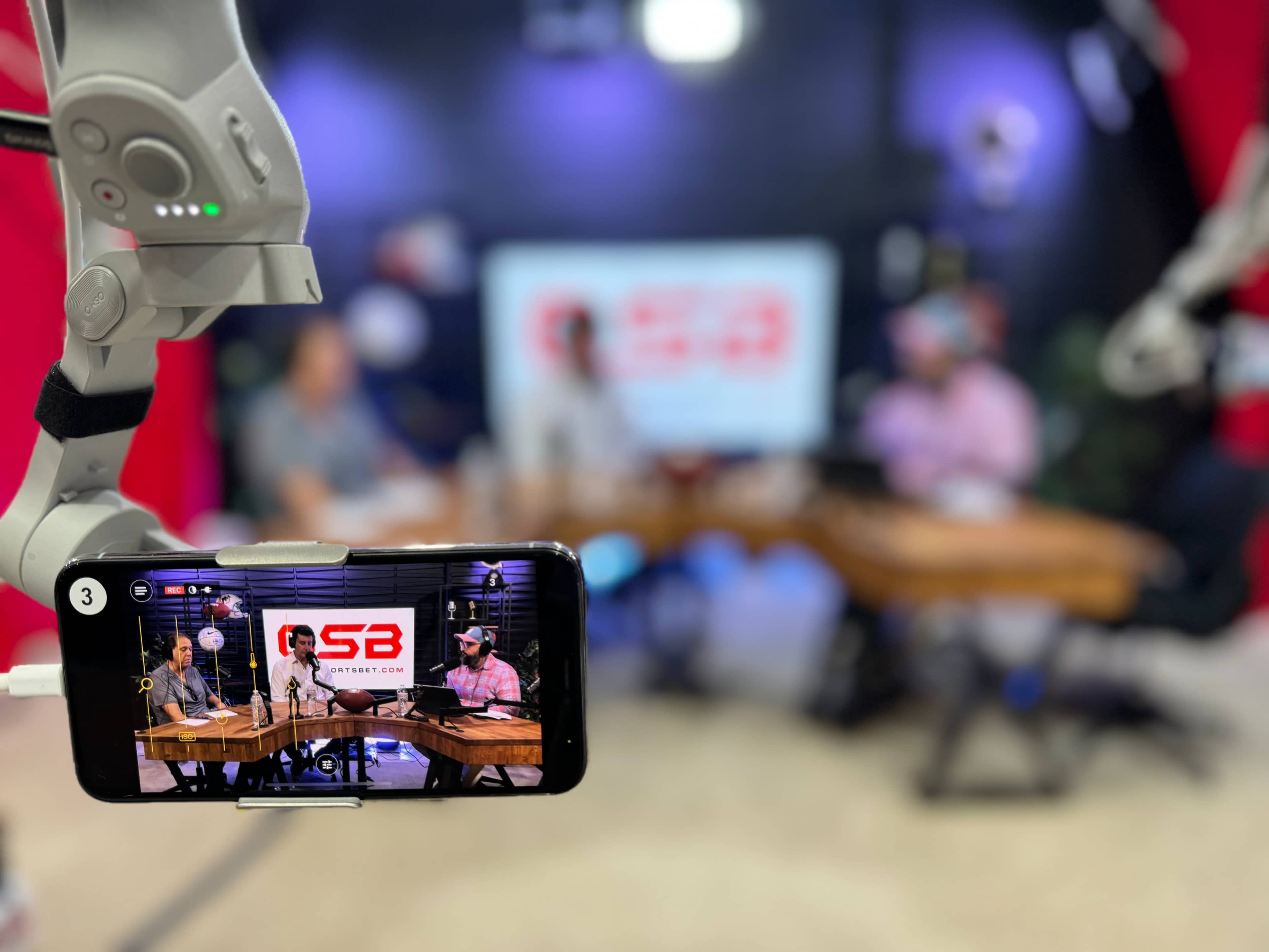 A smartphone on a table captures a live podcast recording featuring several individuals seated around a wooden table in a studio with a large screen displaying a logo.