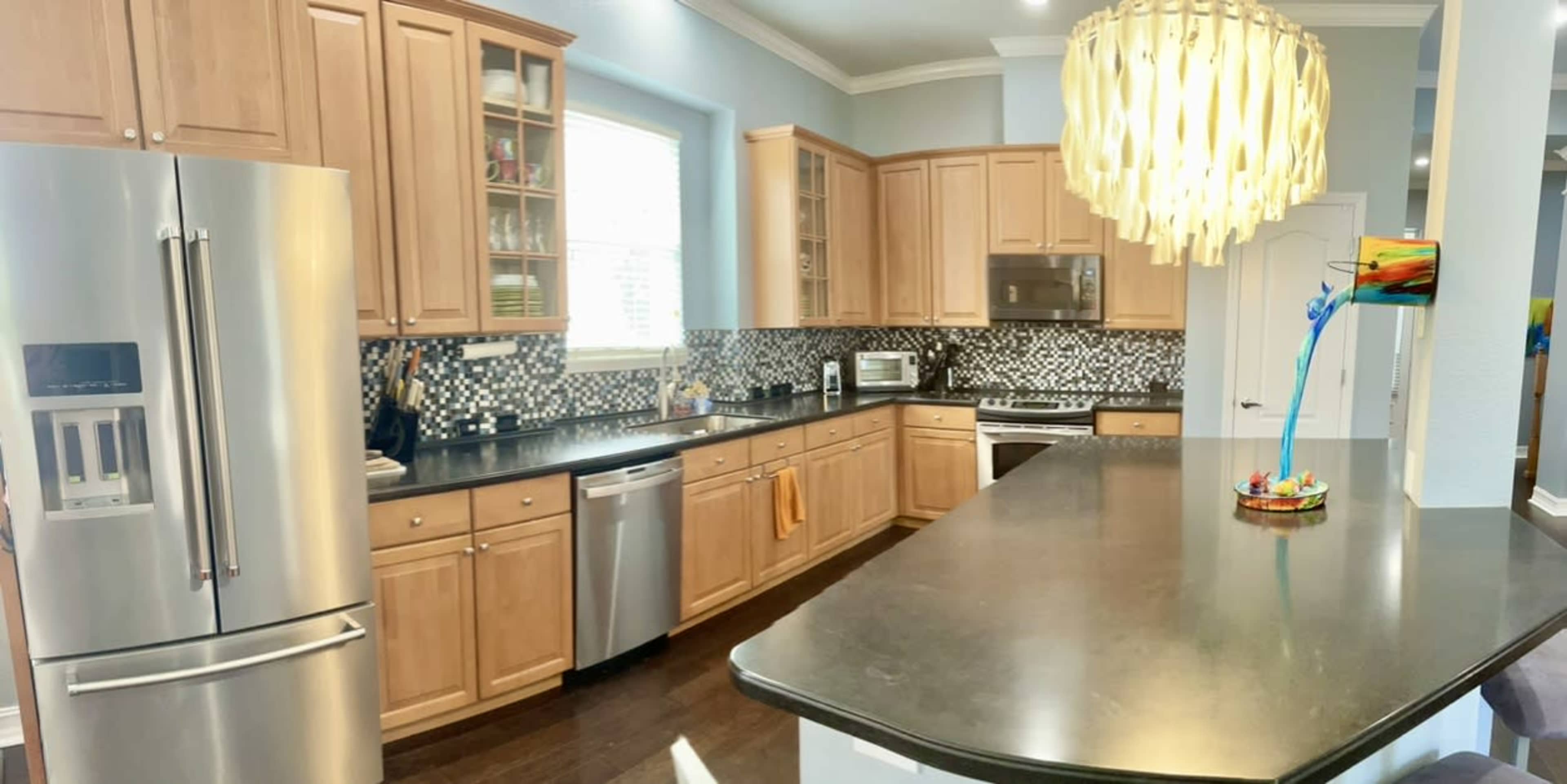 The image shows a modern kitchen with wooden cabinetry, a dark countertop, stainless steel appliances, and a large decorative light fixture above the island.