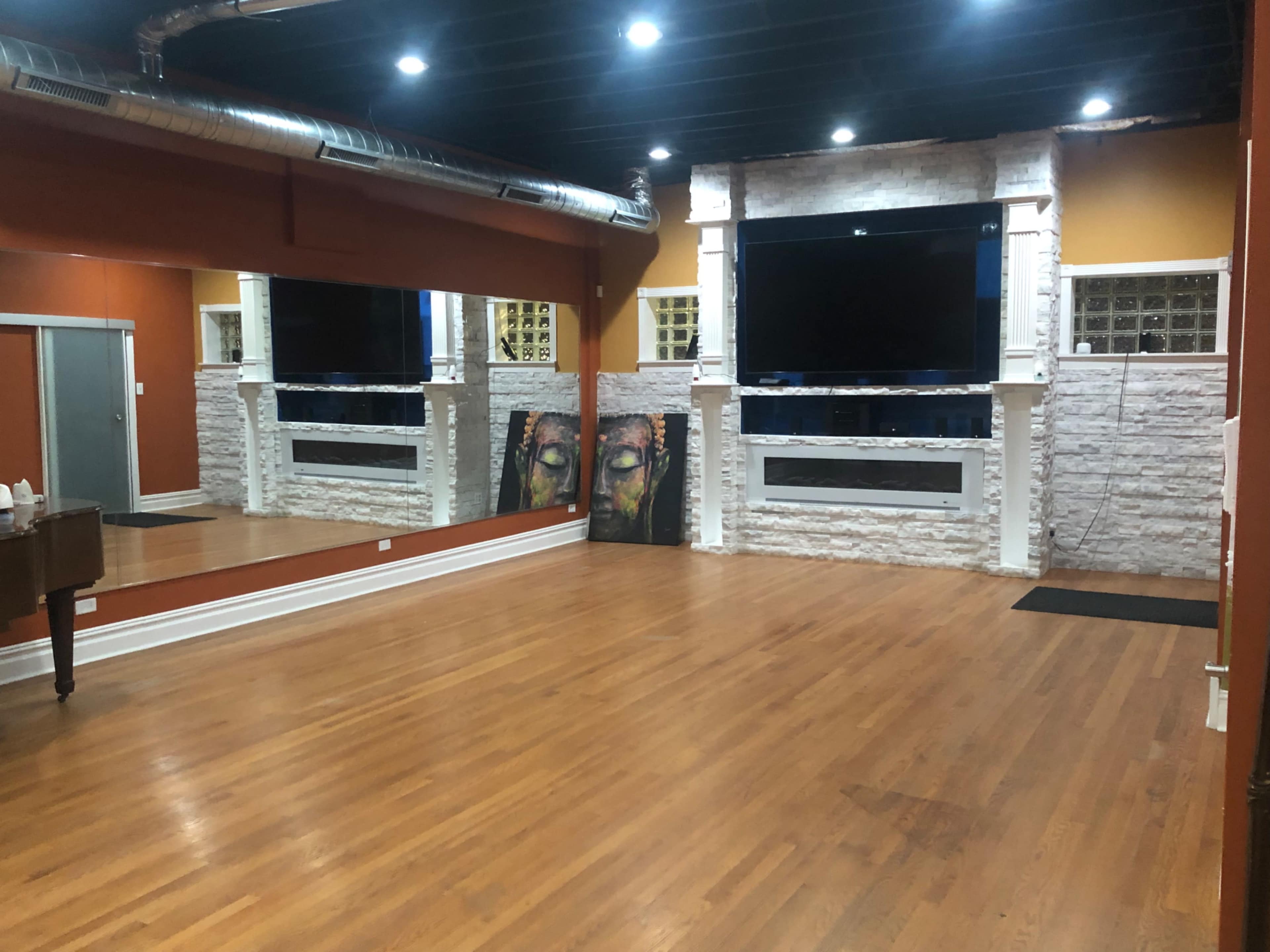 An indoor studio space with wooden floors, a large mirror on one wall, and a prominent flat-screen TV mounted above a white stone feature wall.
