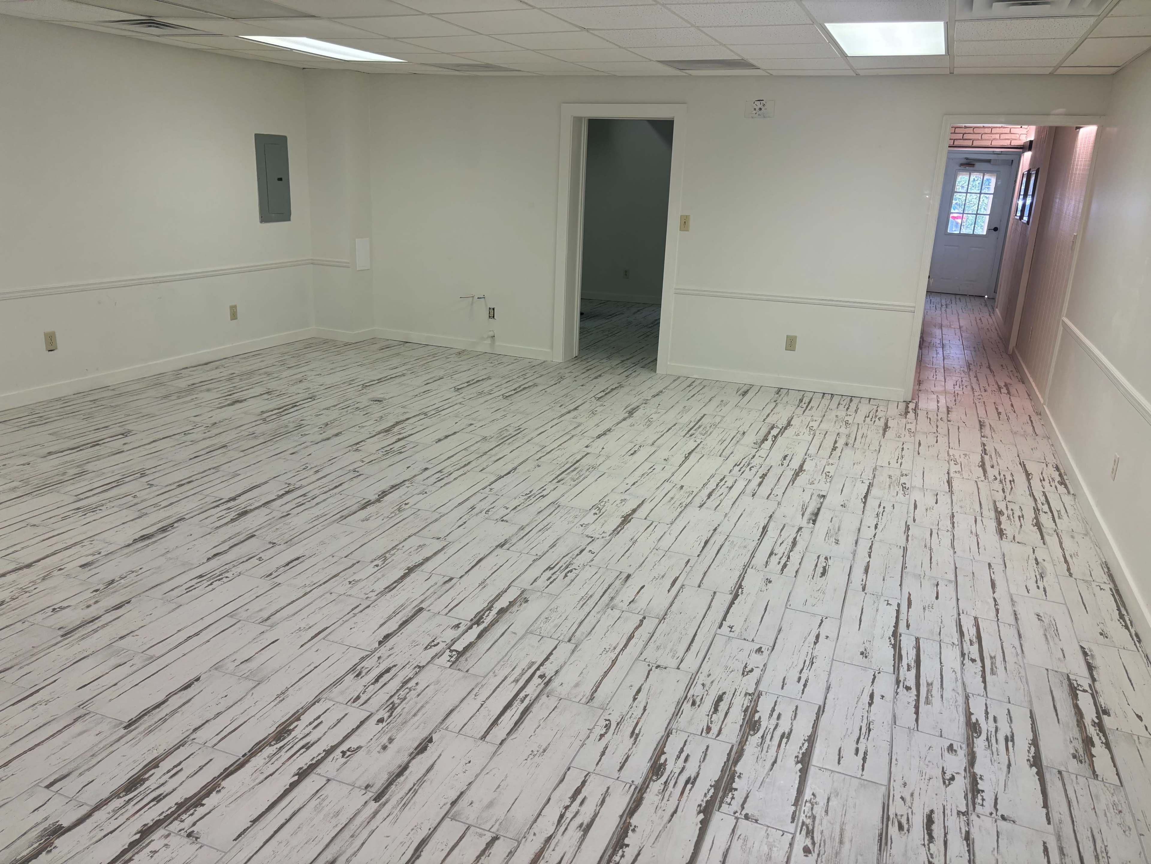 The image shows an empty room with a whitewashed wooden floor and plain white walls, featuring two doorways and a small utility panel on one side.