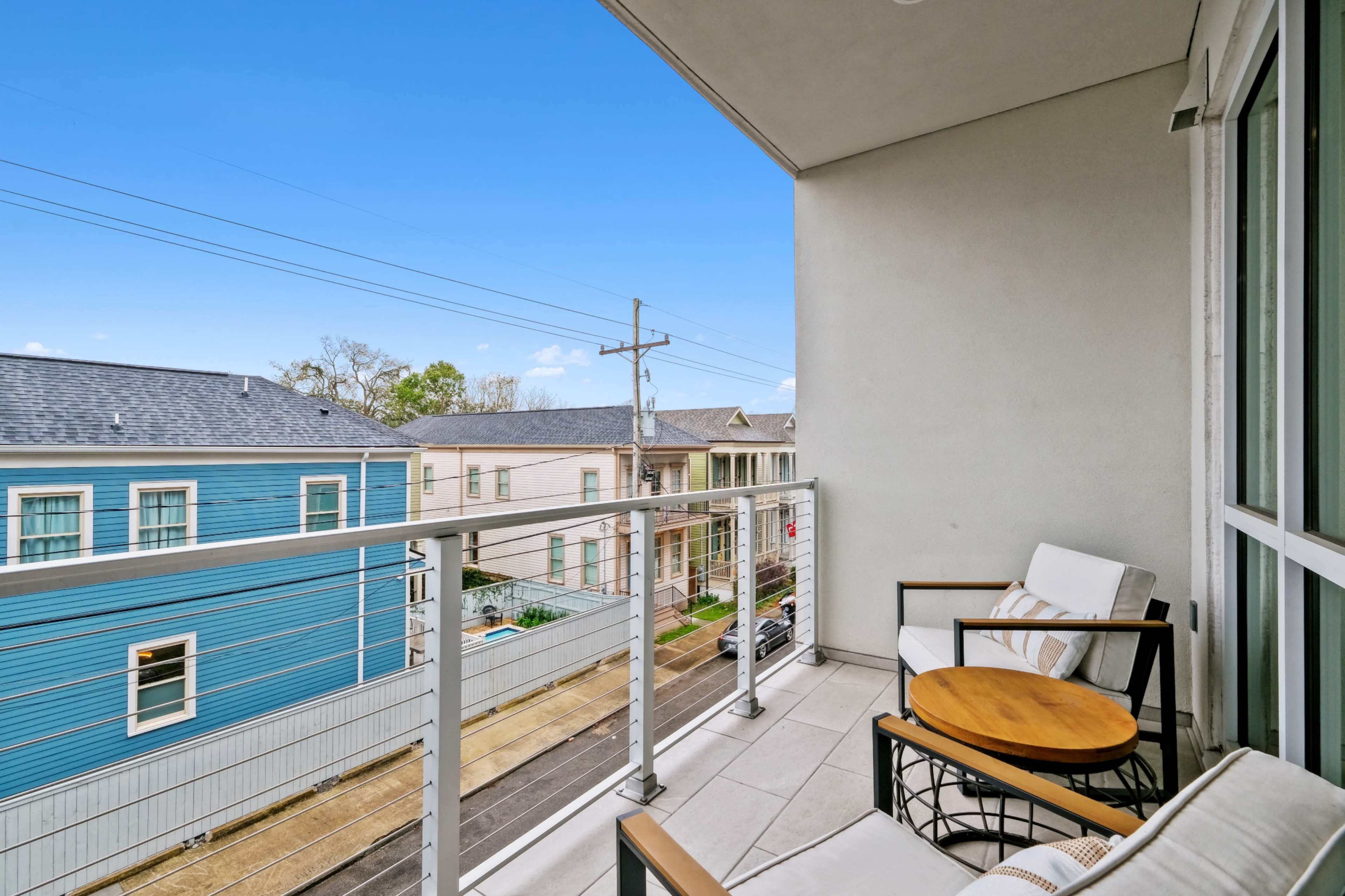 The image shows a balcony with two chairs and a small round table overlooking a street with colorful houses and power lines.