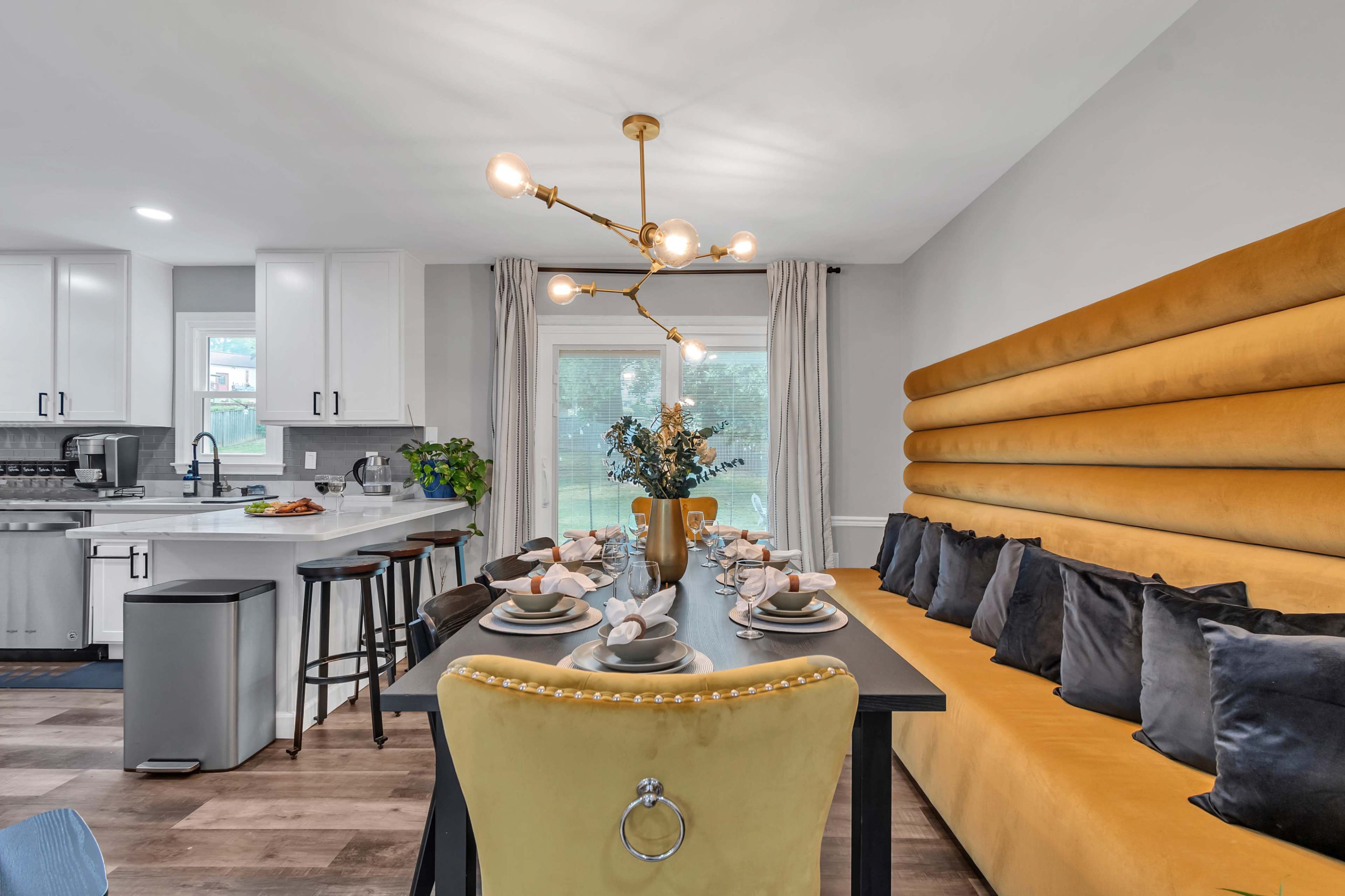 A modern dining area featuring a long table set for a meal, surrounded by a yellow upholstered banquette and stylish chairs.